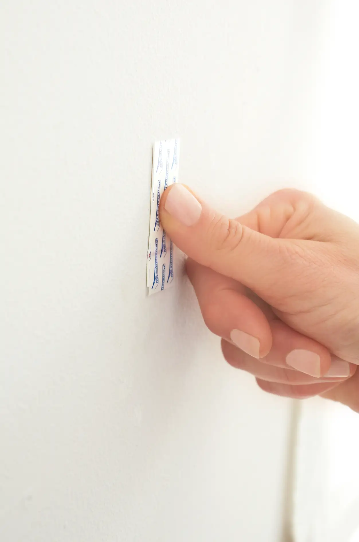 Powerstrip® mindestens fünf Sekunden lang fest auf die gewünschte Stelle an der Wand drücken. Darauf achten, dass die rote Linie des Powerstrips® unten ist. Die Schutzfolie am Anfasser nicht entfernen.