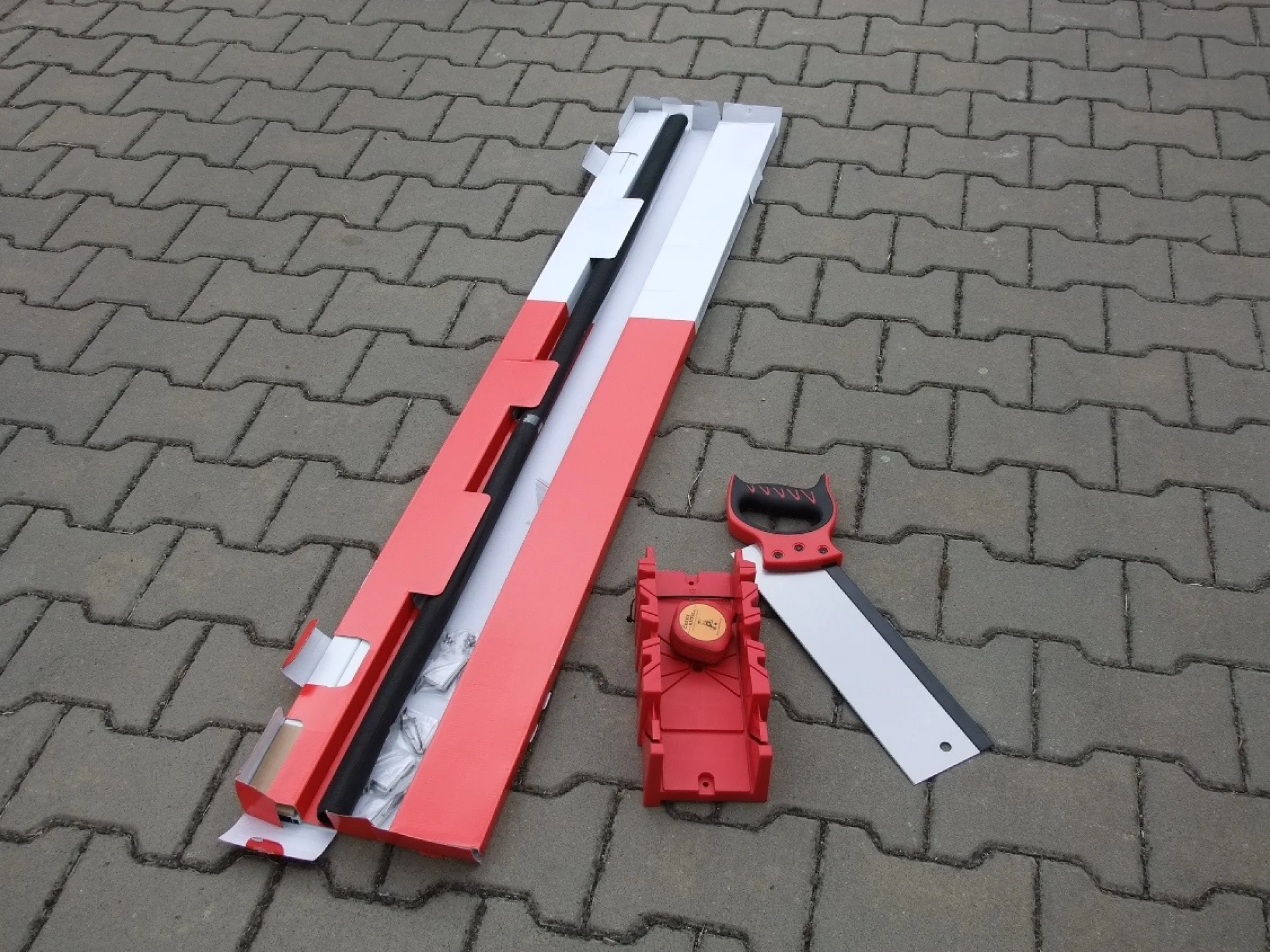 DSCF6589.JPG Červená a černá pila na železo částečně spočívá v otevřené červenobílé krabici na šedém cihlovém chodníku. Viditelná je pila, samostatná červená a černá rukojeť a červený plastový pokosový box s úhloměrem. (This text has been generated by AI)