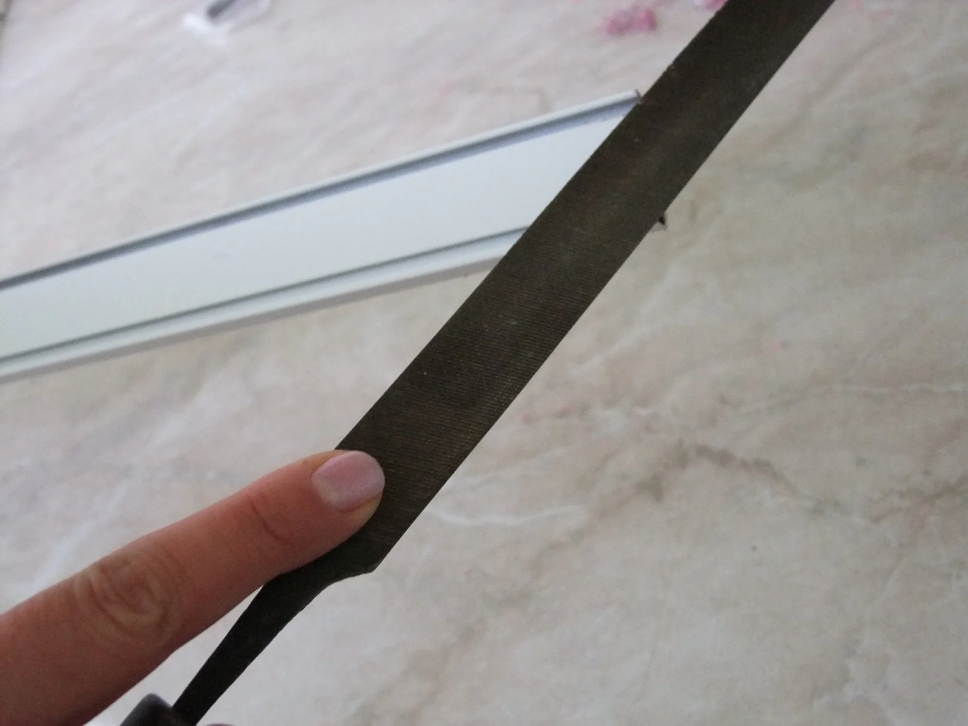 Detail ruky držící plochý kovový pilník na světlém povrchu. Pilník má texturovaný povrch pro vyhlazení nebo tvarování materiálů. Na pozadí je vidět kovový nebo plastový proužek, částečně rozmazaný, s jemně viditelnou páskou tesa. (This text has been generated by AI)