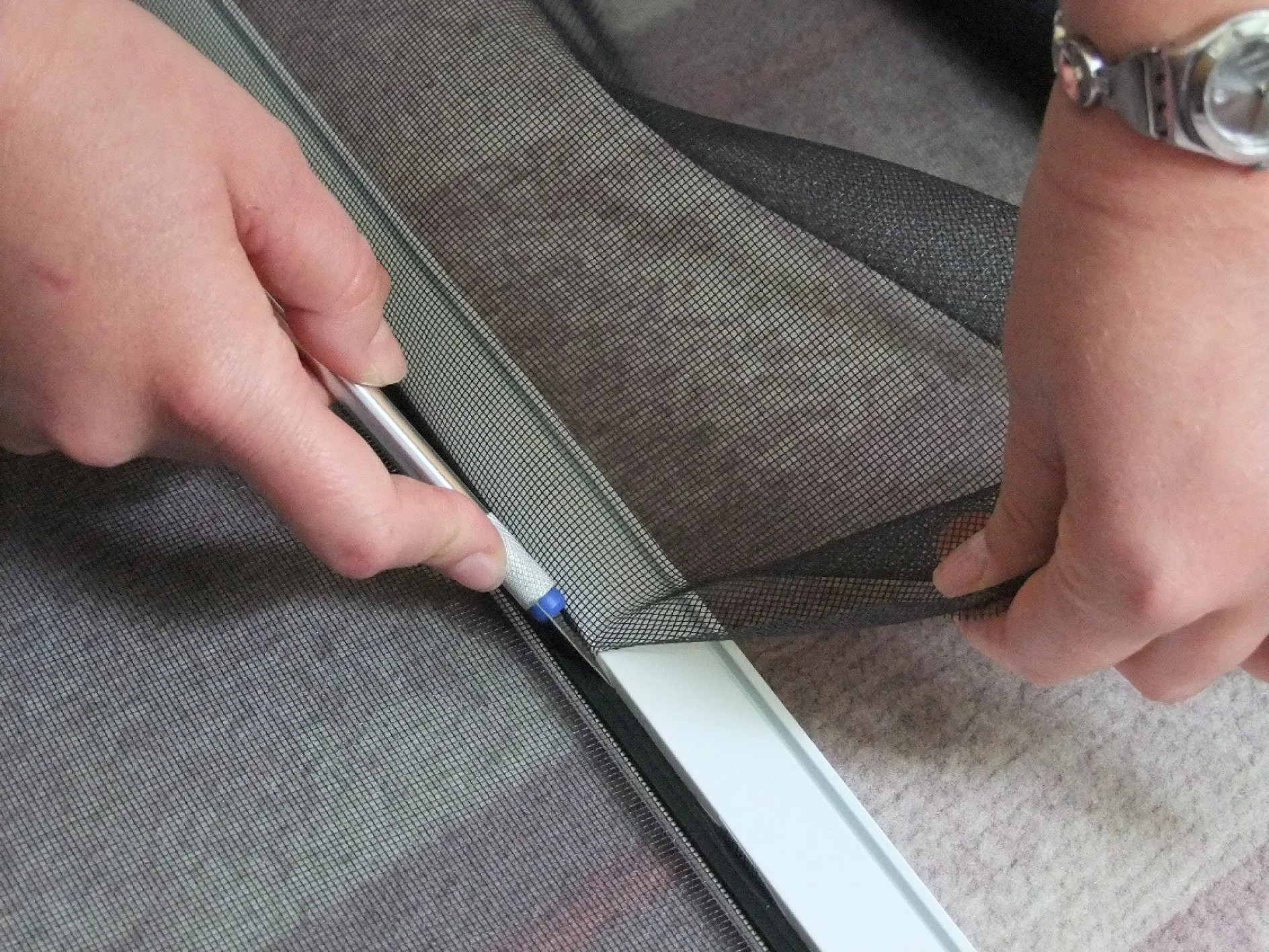 Detail rukou instalují síťovinu do rámu pomocí nástroje. Osoba stříhá nebo fixuje síťovinu páskou tesa s hodinkami na levém zápěstí. Pozadí je neostré. (This text has been generated by AI)