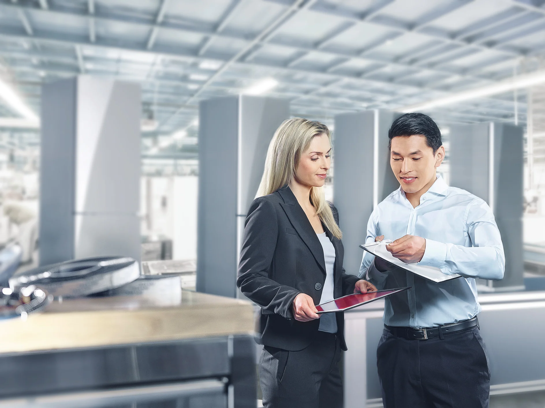 A man and a woman are discussing documents in a bright industrial setting. The woman holds a tablet, and the man holds a clipboard secured with tesa tape. They are both professionally dressed. Equipment and large metal structures are visible in the background. (This text has been generated by AI)