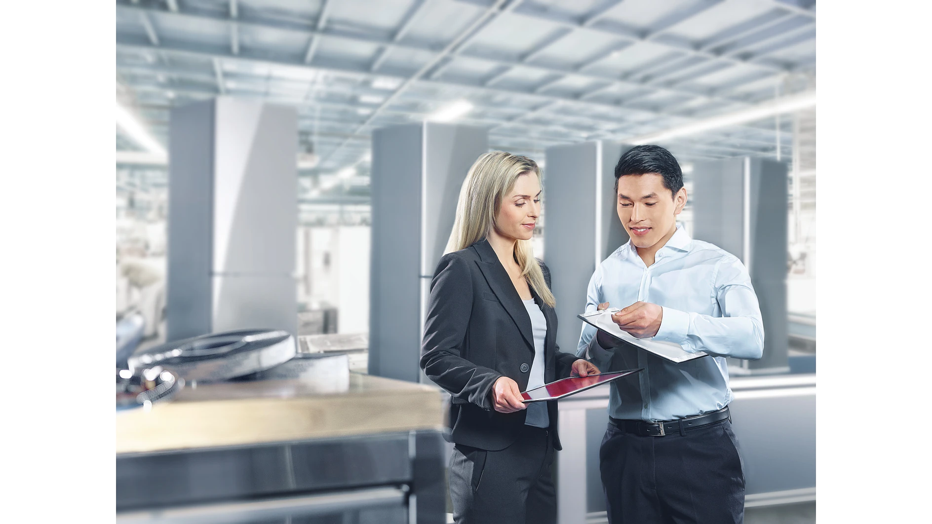 A man and a woman are discussing documents in a bright industrial setting. The woman holds a tablet, and the man holds a clipboard secured with tesa tape. They are both professionally dressed. Equipment and large metal structures are visible in the background. (This text has been generated by AI)