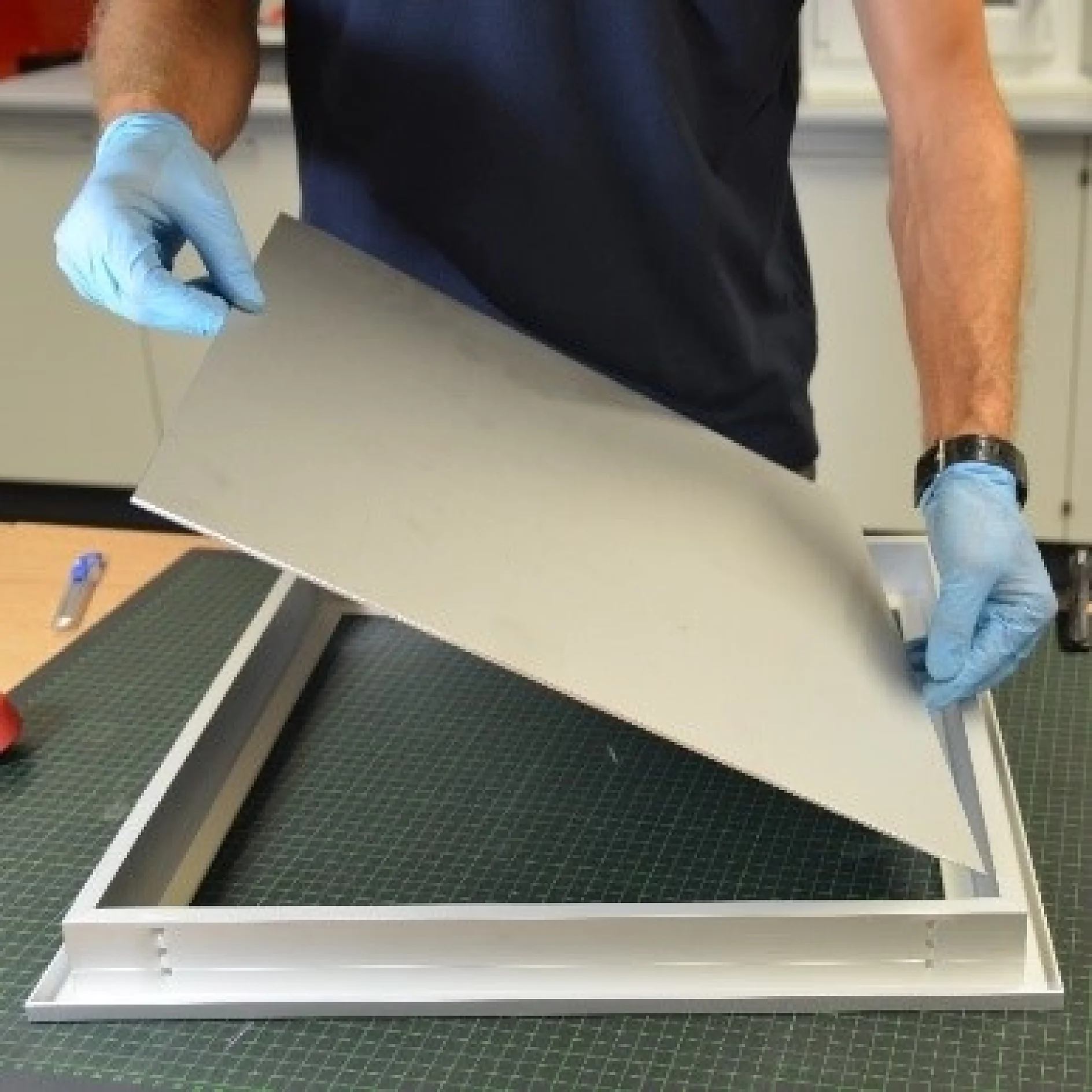 A person wearing a dark short-sleeve shirt and blue gloves is assembling or disassembling a metal frame and panel. They are holding a rectangular metal sheet above a rectangular metal frame on a green grid mat, using tesa tape for secure positioning. (This text has been generated by AI)