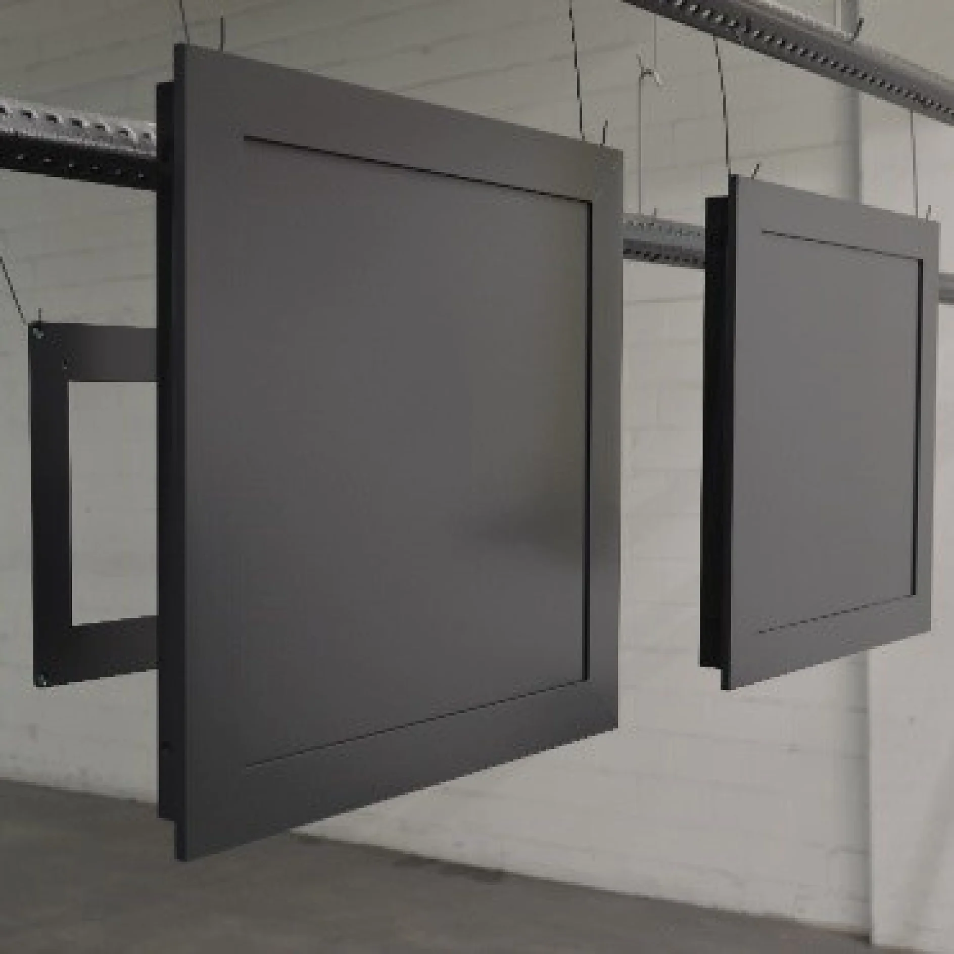 Two black, square metal frames hang from a rail inside an industrial-looking space with a white brick wall. The frames are suspended at different angles using tesa tape, suggesting they are part of a manufacturing process. (This text has been generated by AI)