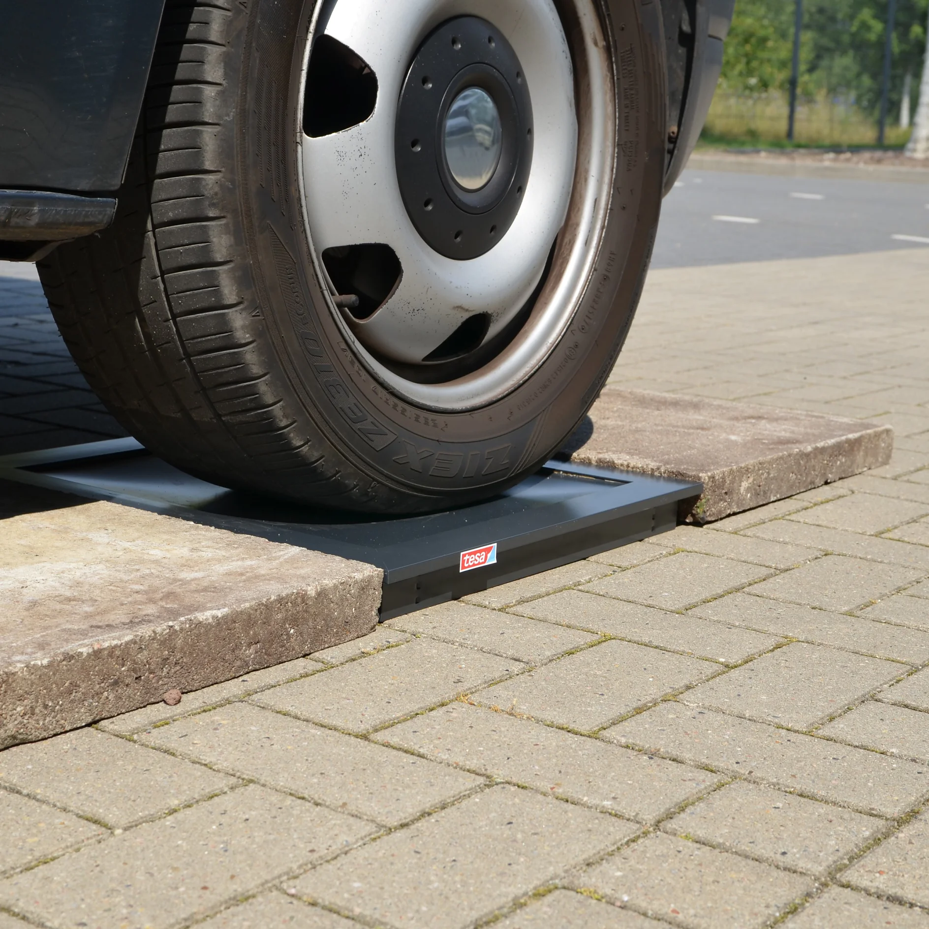 A car tire is positioned on a black metal platform placed on a paved area, secured with tesa tape. The platform is flanked by two concrete slabs. The setting appears to be outdoor, with a portion of the road visible in the background. (This text has been generated by AI)