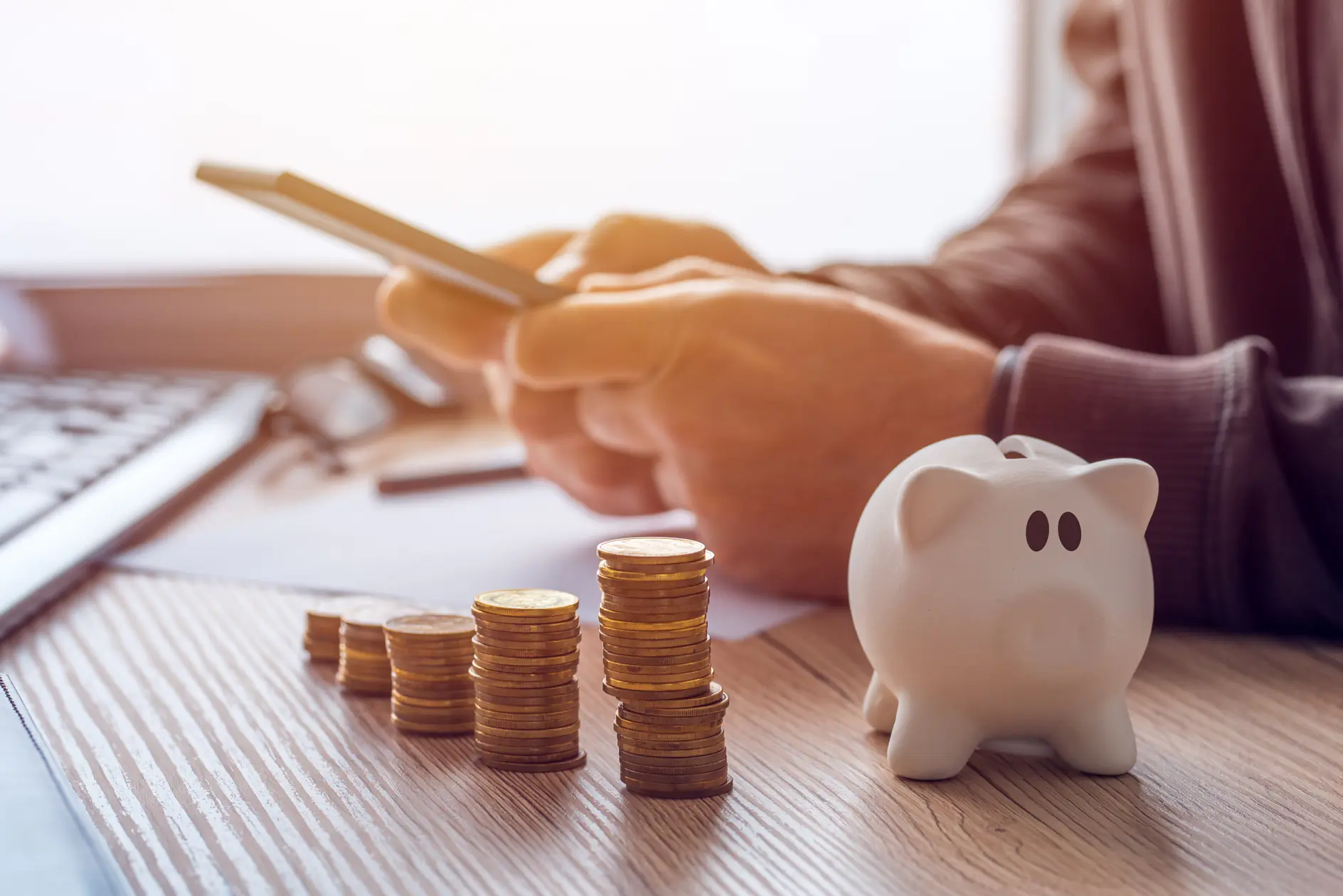 A person uses a smartphone at a desk with neatly stacked coins and a white piggy bank. The scene suggests financial planning or online banking, with a blurred keyboard and papers visible in the background. (This text has been generated by AI)
