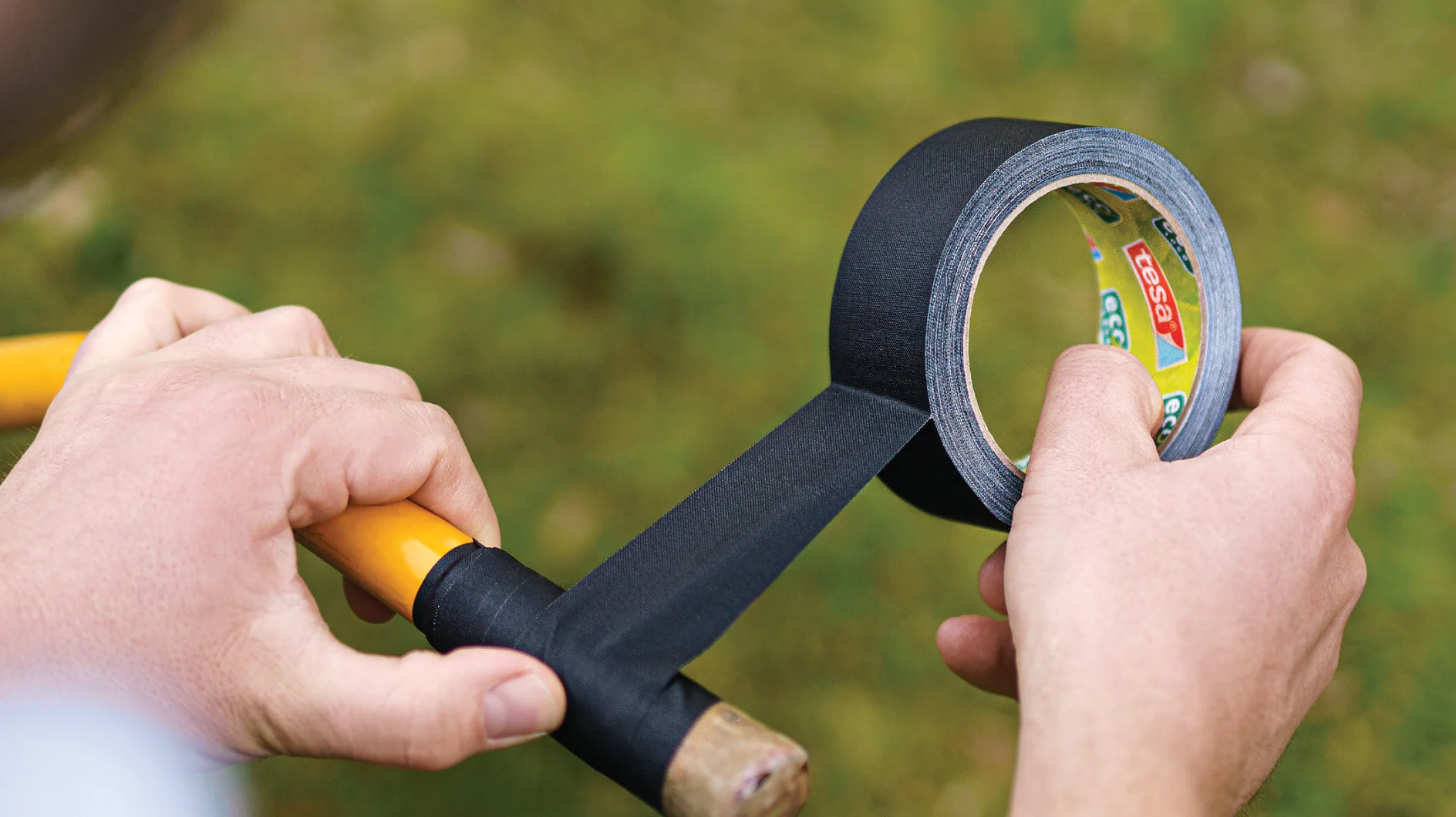 A person wraps black tesa tape around the handle of a yellow tool. The tesa tape is being unrolled from a branded roll. The background is blurred with green and brown tones, suggesting an outdoor setting. (This text has been generated by AI)