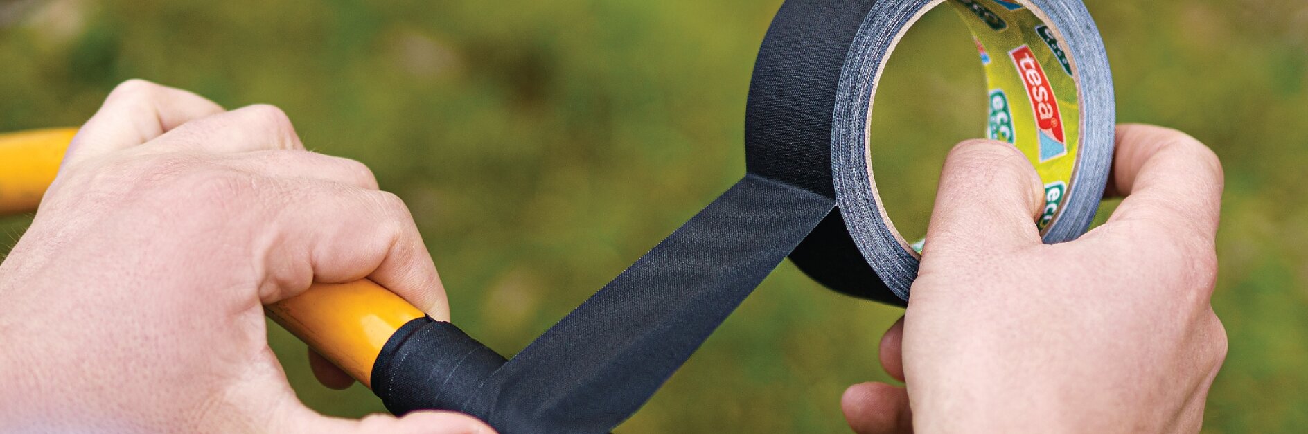 A person wraps black tesa tape around the handle of a yellow tool. The tesa tape is being unrolled from a branded roll. The background is blurred with green and brown tones, suggesting an outdoor setting. (This text has been generated by AI)