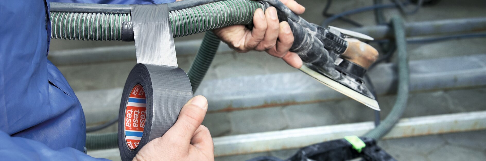 A person in a blue jacket is using silver tesa tape to secure a green ribbed hose to a metal tool with a rectangular base. The tool is connected to a piece of equipment with wheels in the background.