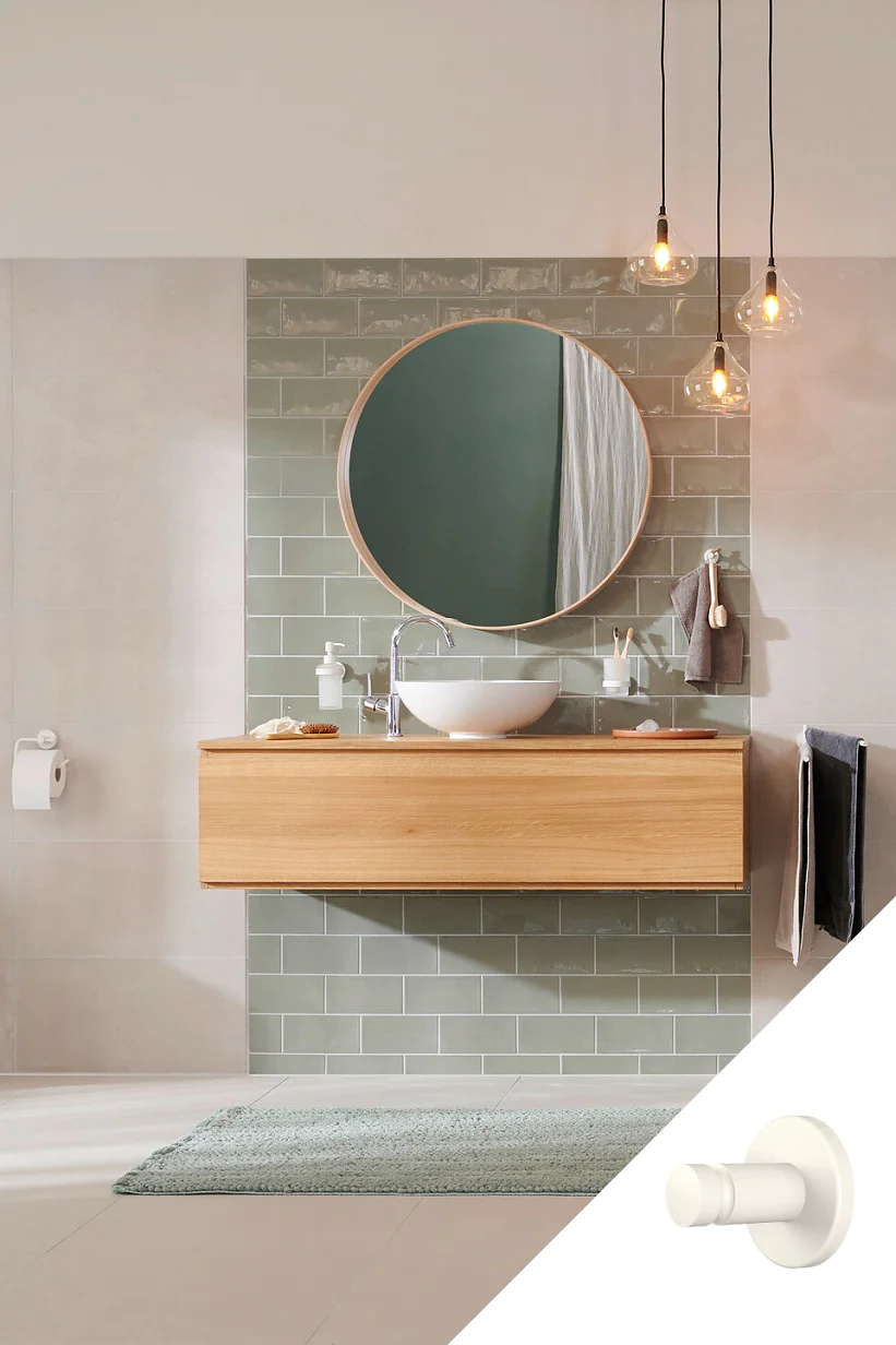 Minimalistická koupelna s kruhovým zrcadlem nad dřevěným umyvadlem s bílým umyvadlem a baterií. Zelené dlaždice metra tvoří backsplash. Světlo zajišťují tři závěsné Edisonovy žárovky. Ručník je na stojanu, zajištěný páskou tesa a na podlaze leží modrý koberec. (This text has been generated by AI)