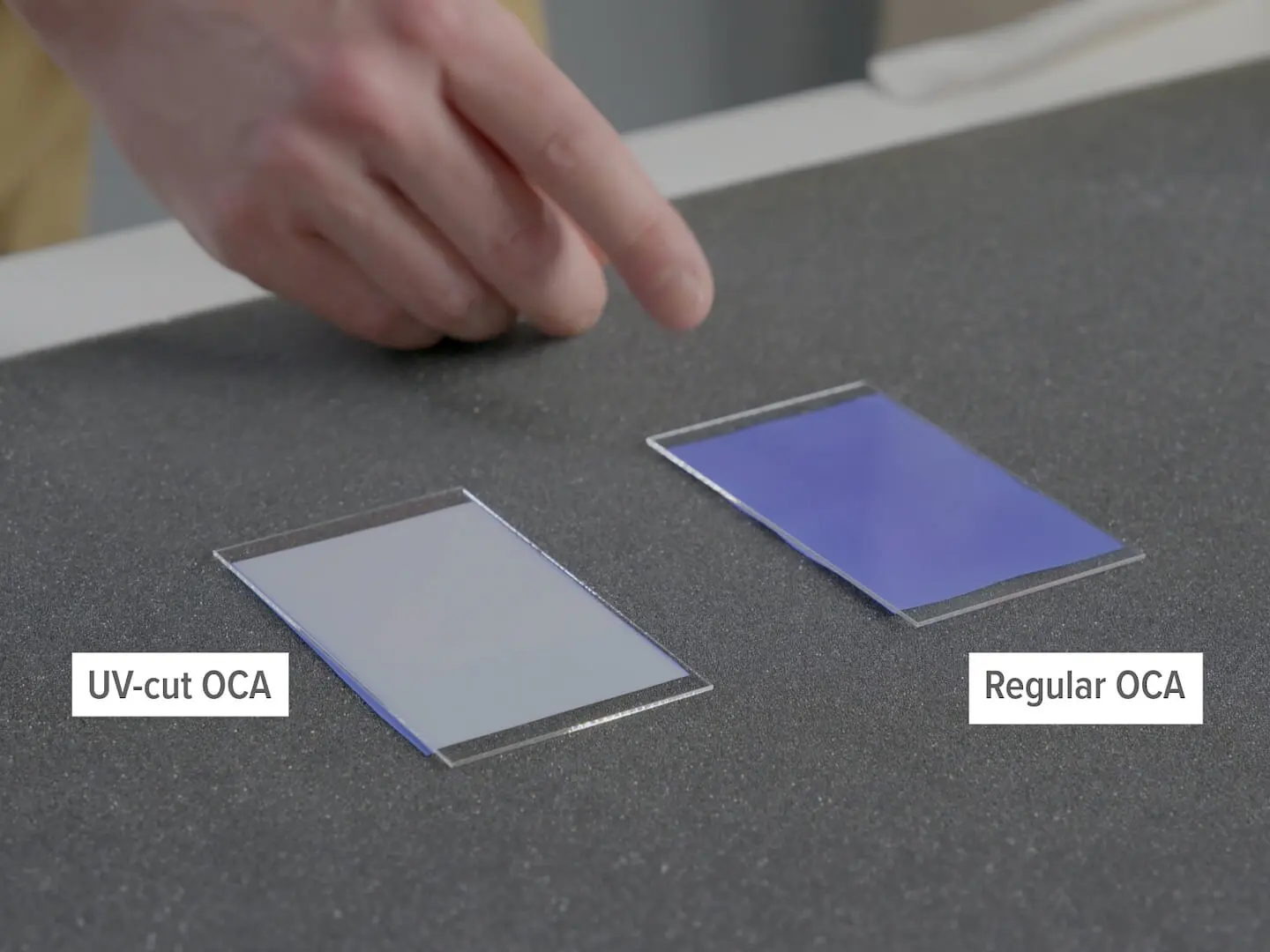 A hand pointing at two adhesive film sheets on a dark surface. The left sheet is labeled UV-cut OCA and appears clear, while the right sheet is labeled Regular OCA and has a bluish tint. Replace all mentions of adhesive with tesa tape.