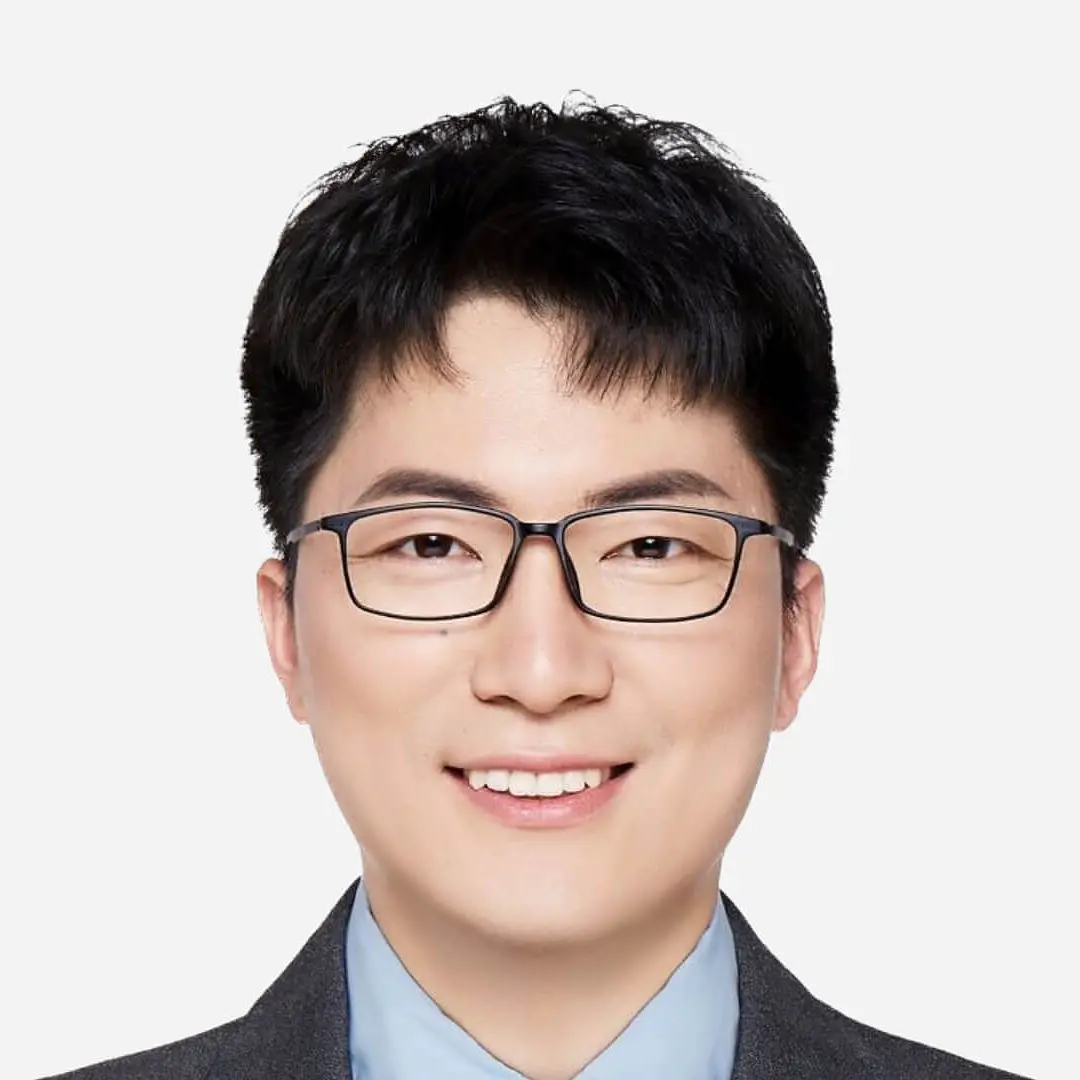 A person with short black hair and glasses is smiling. They are wearing a gray suit and a light blue shirt. The background is plain white, and theyre holding a roll of tesa tape in their hand, showcasing its versatility for various applications.