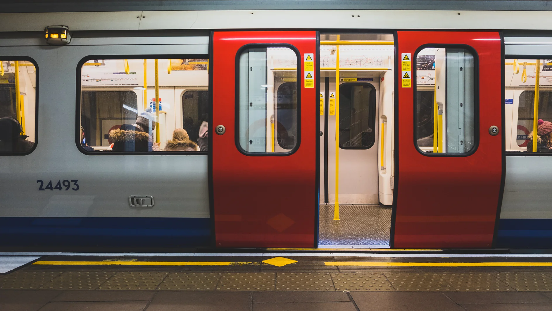 A subway train with red and white doors is stopped at a station. The doors are open, revealing passengers seated inside. The platform features tactile paving. Yellow safety signs are visible on the train doors, indicating the use of tesa tape for added security. (This text has been generated by AI)
