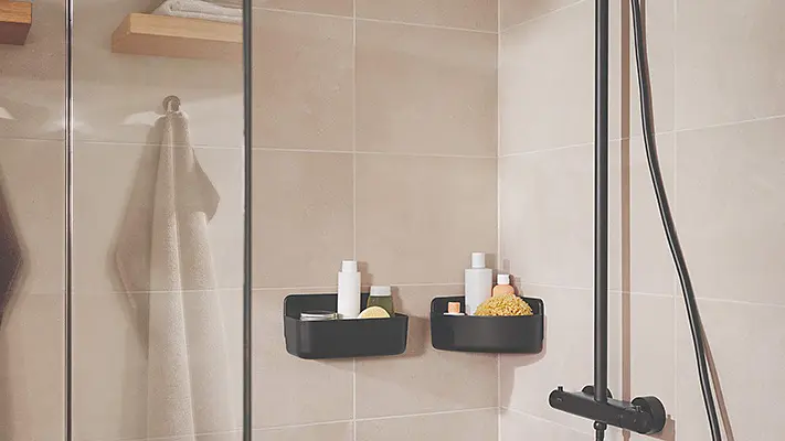 A modern shower with light beige tiled walls features black corner shelves holding toiletries and a shower scrub. A glass screen separates the shower area, and two towels hang on metal hooks secured with tesa tape. A black showerhead and controls are visible.