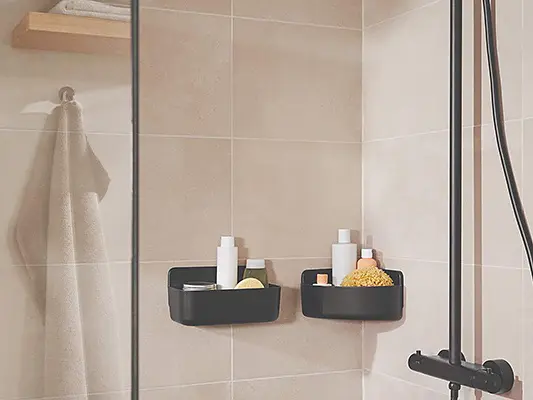 A modern shower with light beige tiled walls features black corner shelves holding toiletries and a shower scrub. A glass screen separates the shower area, and two towels hang on metal hooks secured with tesa tape. A black showerhead and controls are visible.