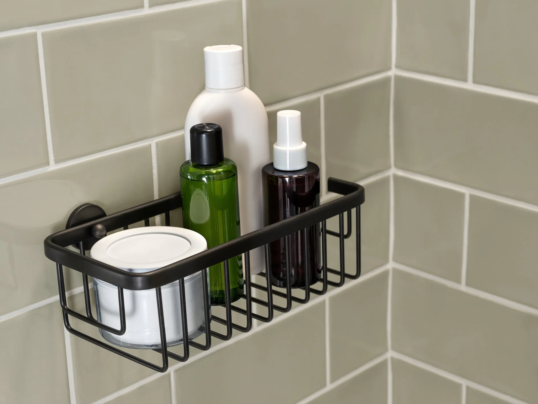 A shower caddy mounted on a tiled wall holds various toiletries, including a white bottle, a green bottle, a spray bottle, and a small white container. The tiles are a light grayish-green color, and the caddy is securely attached with tesa tape. (This text has been generated by AI)