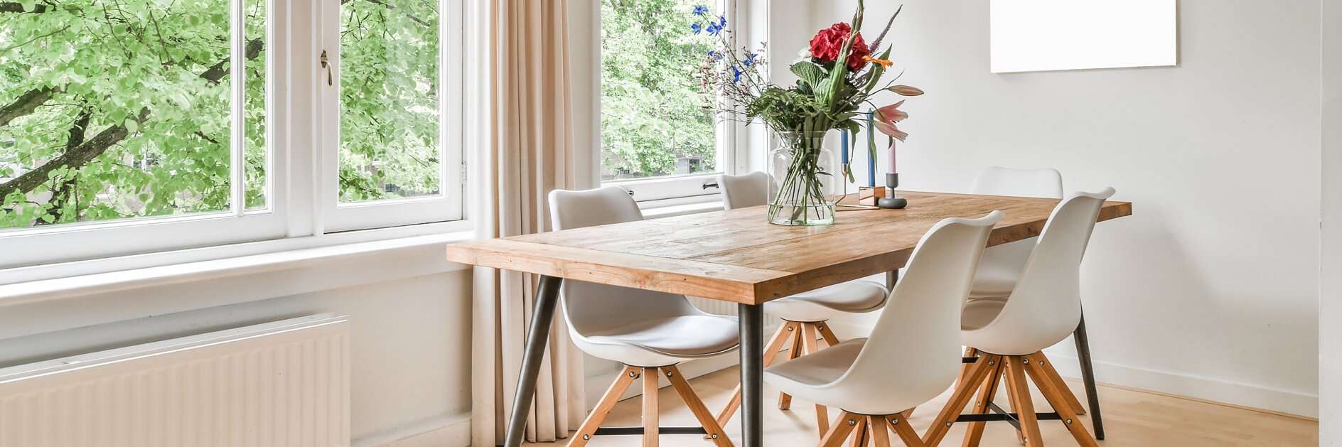 A bright dining room features a wooden table accompanied by six white chairs. On the table sits a vase filled with flowers. To the left, large windows offer a view of green trees. The decor includes a single hanging light and an empty frame on the wall. The floor is crafted from wood. (This text has been generated by AI)
