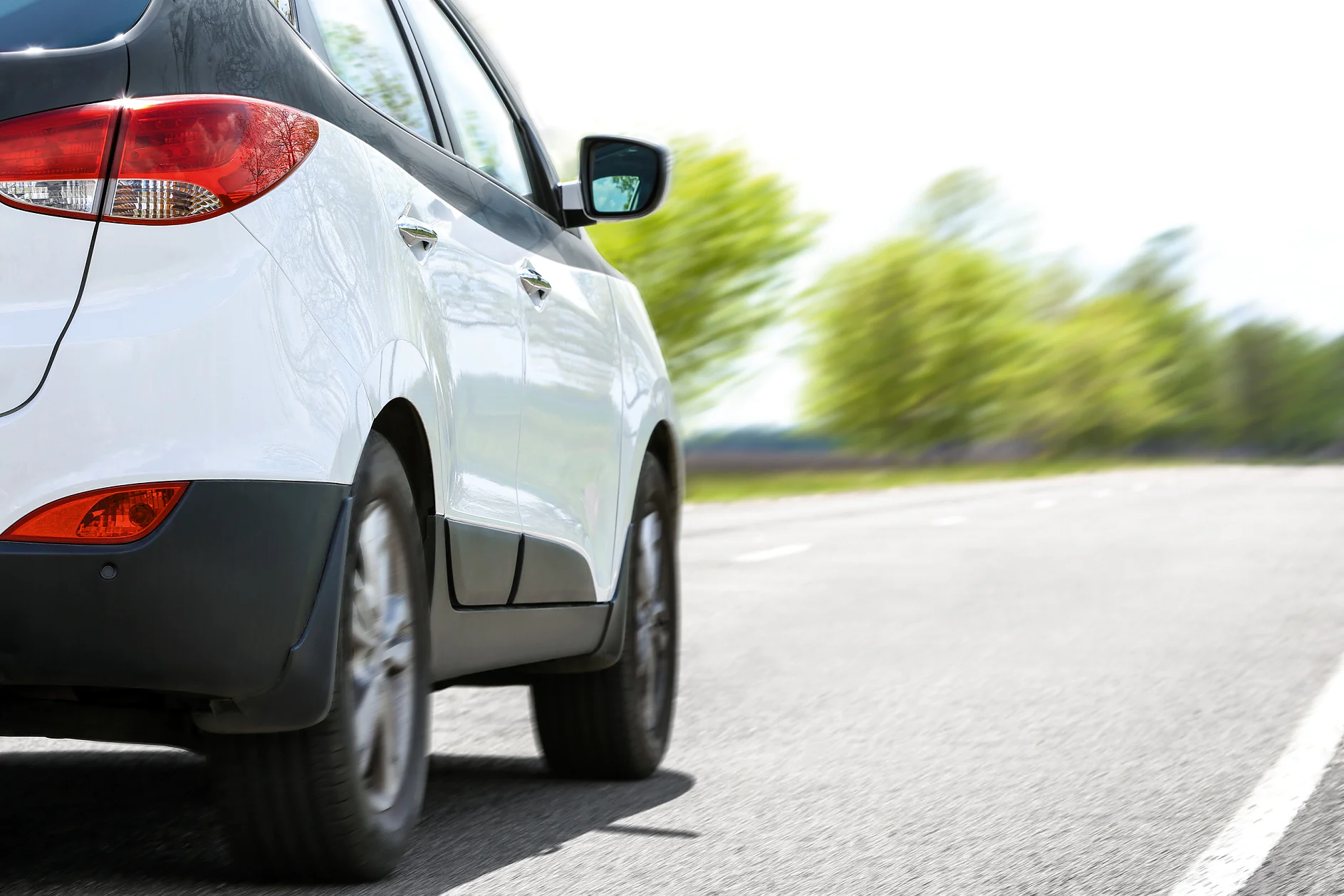 white compact car driving on a paved road with blurred green trees in the background on a clear day (This text has been generated by AI)