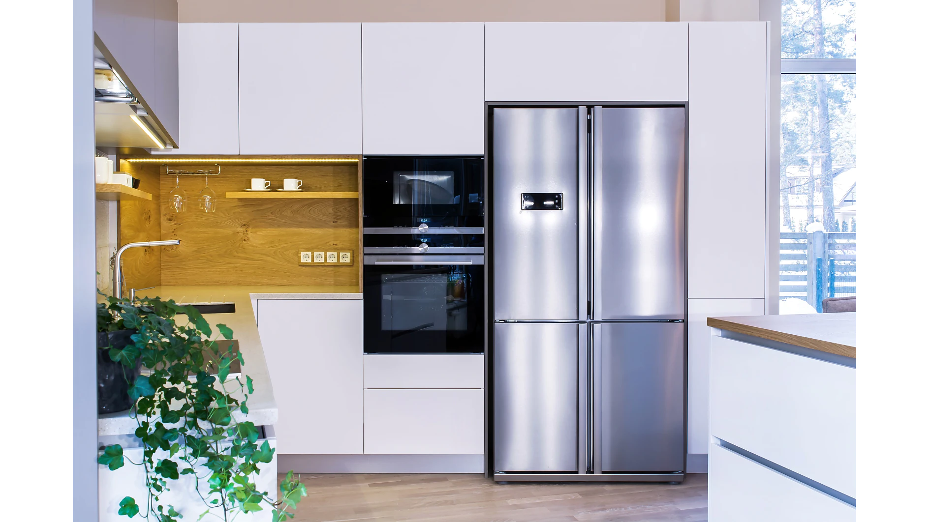 Moderní kuchyně s bílými skříňkami, vestavěnou černou troubou a mikrovlnnou troubou, dřevěným obkladem s otevřenými policemi zajištěnými páskou tesa a velkou nerezovou dvoukřídlou lednicí. Na pracovní desce je vidět zelená rostlina. (This text has been generated by AI)