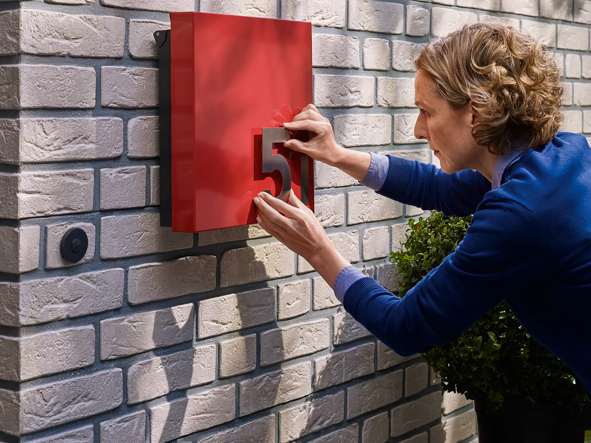 Person affixing the number 5 to a red box mounted on a light brick wall using tesa tape. There is a small round doorbell button nearby, and a potted plant is partially visible. The person is wearing a blue sweater. (This text has been generated by AI)