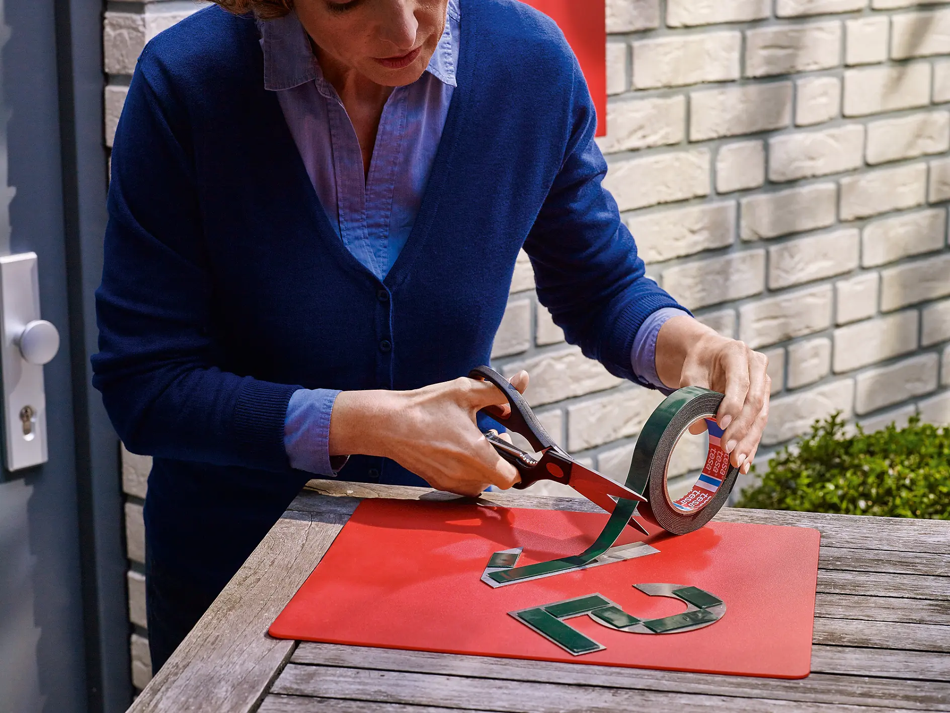 A person cuts a strip of adhesive tesa tape with scissors next to shiny, metallic house numbers 7 and 2 on a red mat placed on a wooden table. The person is wearing a blue sweater over a light blue shirt. The background has a brick wall. (This text has been generated by AI)