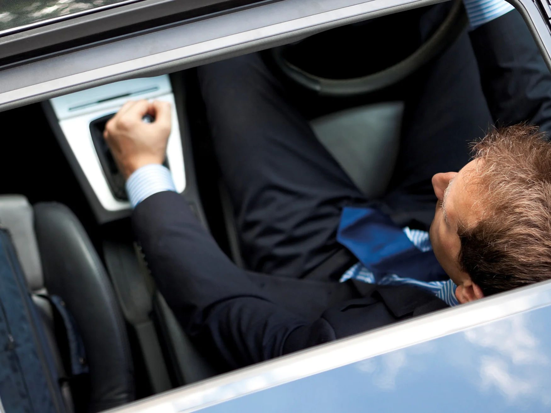 A man in a suit is seated inside a car, viewed from above through an open sunroof. He is adjusting a control on the cars dashboard. The interior appears contemporary, and a steering wheel is visible. (This text has been generated by AI)