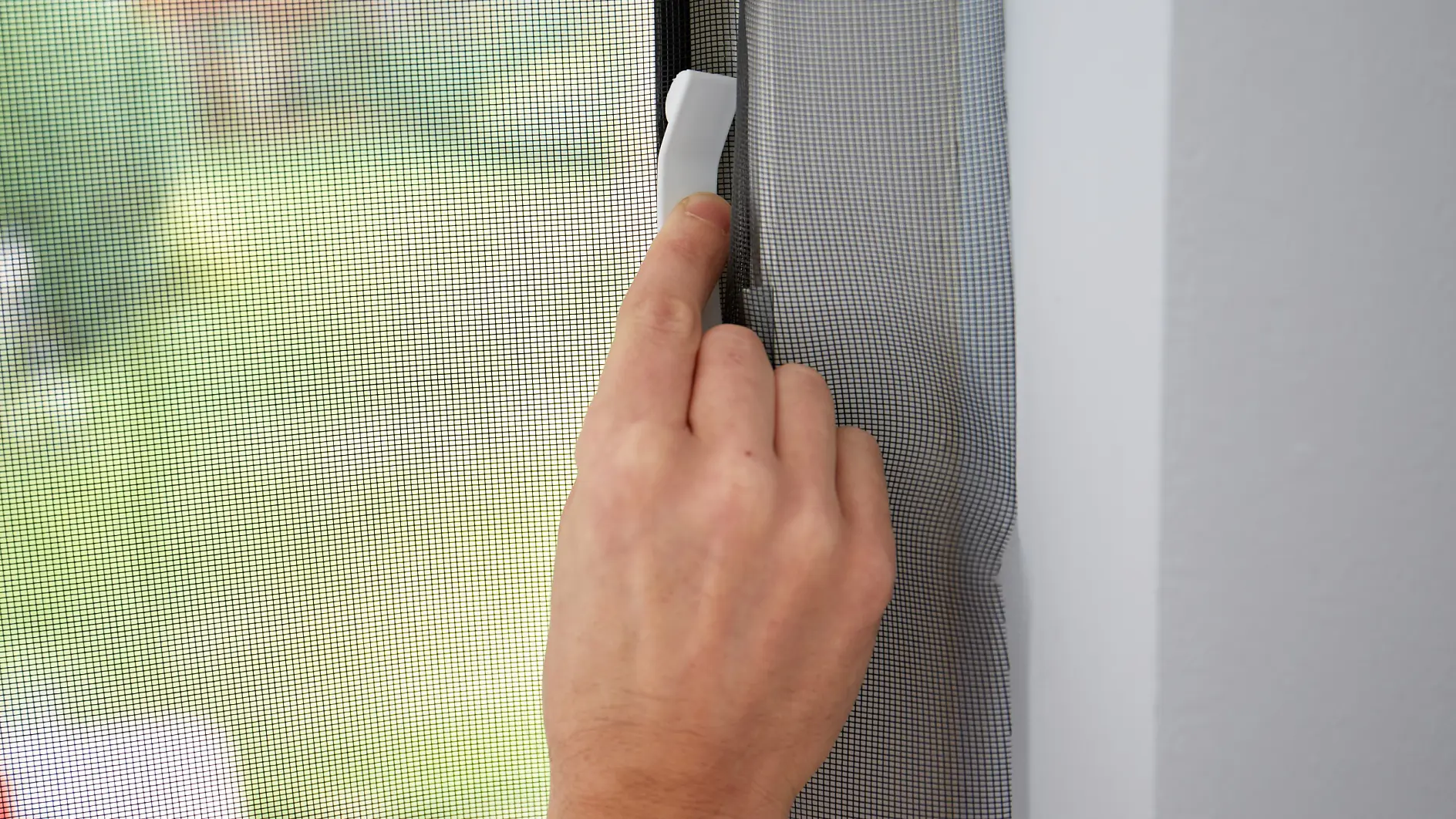A hand is seen opening a retractable window screen using a white handle. The background shows an out-of-focus outdoor scene through the mesh screen secured with tesa tape. (This text has been generated by AI)