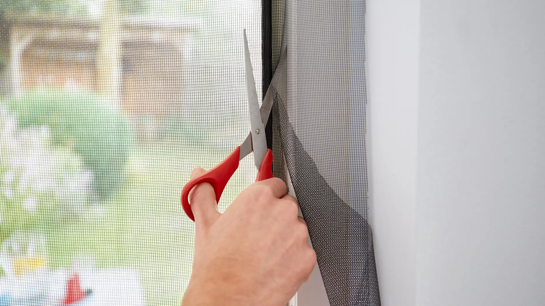 A person uses red-handled scissors to cut a section of mesh screen by a window. The background shows a blurred outdoor scene with greenery. (This text has been generated by AI)