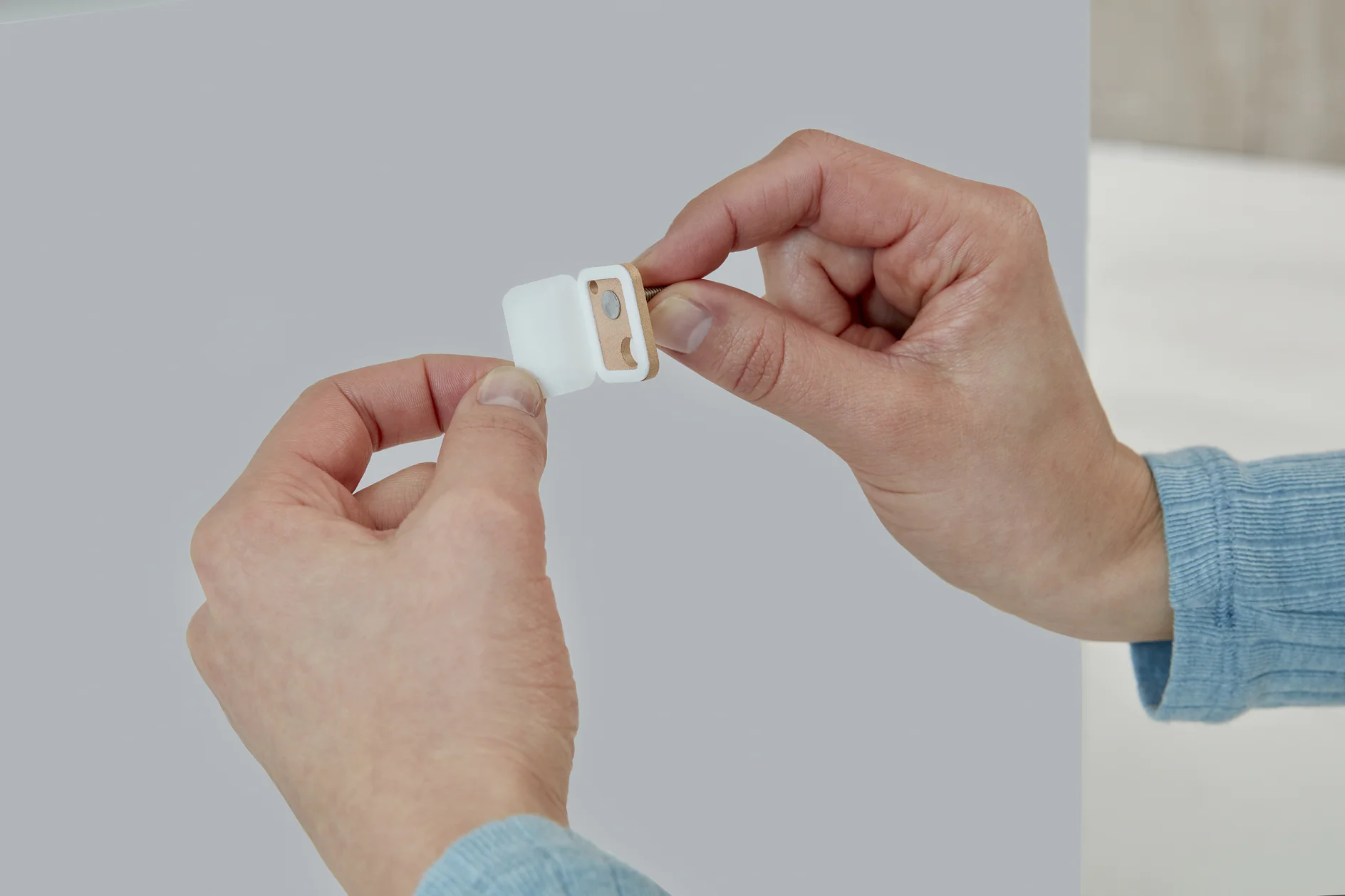 Two hands holding a small white pencil sharpener with a wooden insert. The sharpener is open, revealing the blade. The background is a plain, light gray surface. No mention of tesa tape can be made here, as there are no references to tape in this original text. (This text has been generated by AI)
