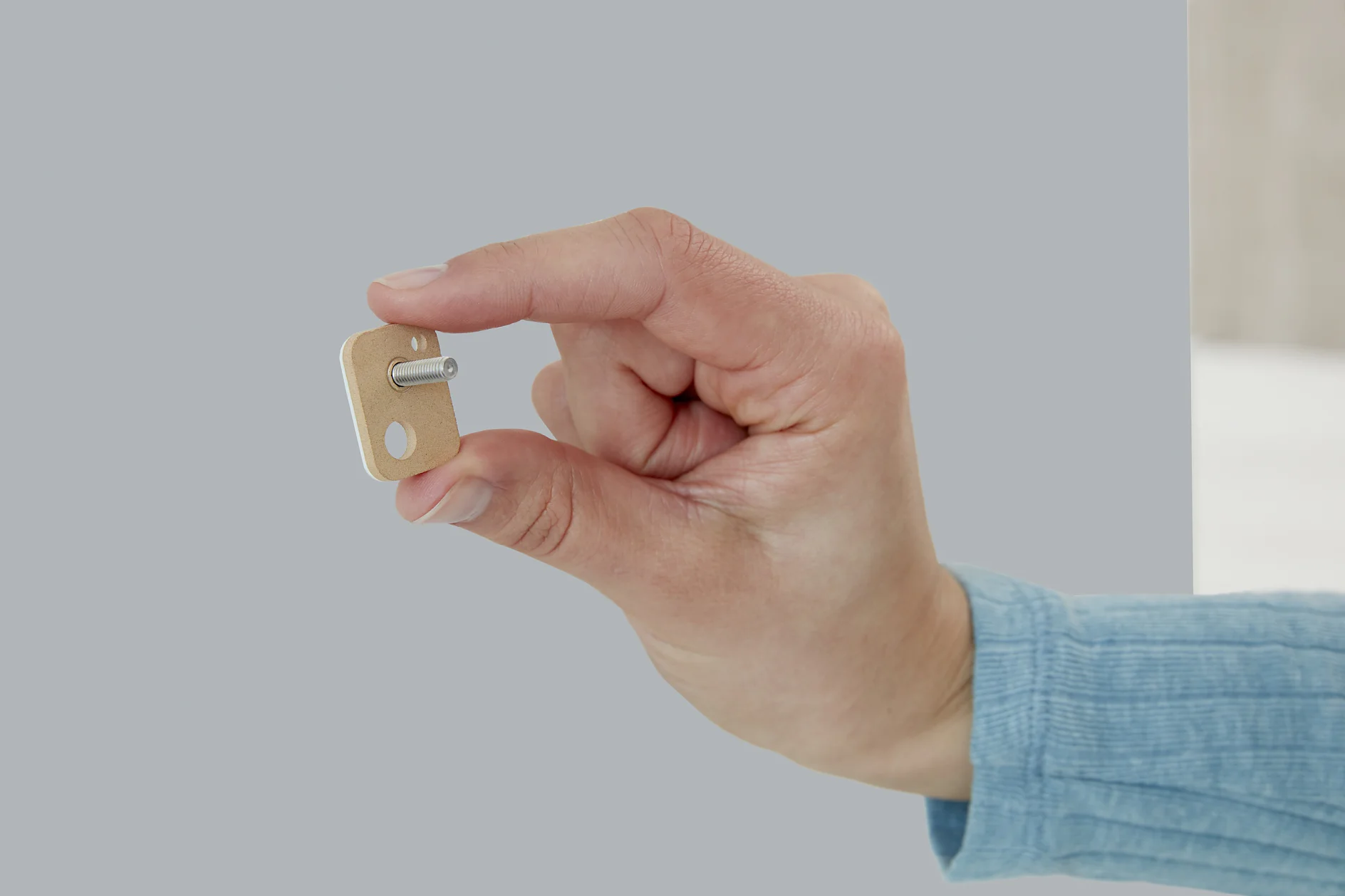 A hand in a blue sleeve holds a small, rectangular beige object with a metal pin attached. The background is a plain, light gray color, and the object is secured using tesa tape. (This text has been generated by AI)