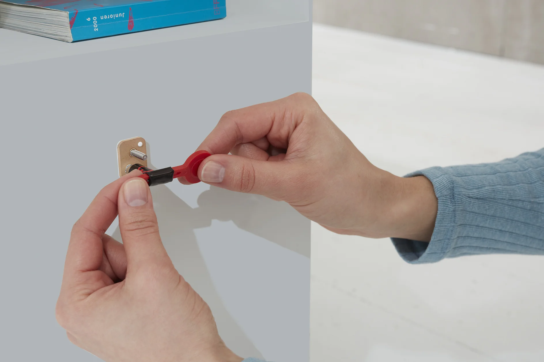 A person wearing a blue long-sleeve shirt is inserting a small screwdriver into a brass hinge attached to a light gray cabinet using tesa tape. A stack of blue books is visible on top of the cabinet. (This text has been generated by AI)