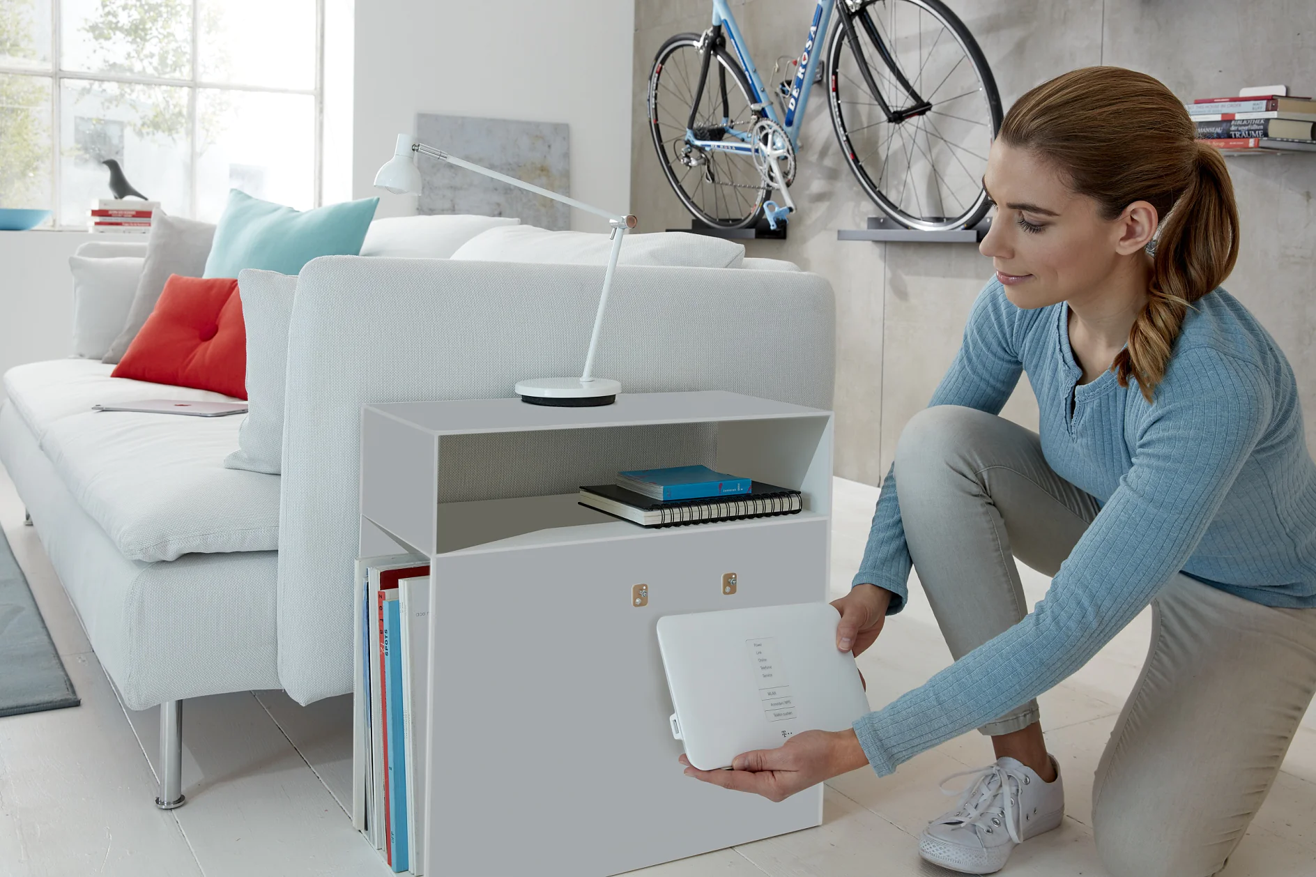 A woman in a light blue sweater kneels beside a white cabinet with open shelving, placing or removing a Wi-Fi router. A sofa, books, a desk lamp, and a mounted bicycle are visible in the modern room. (This text has been generated by AI)