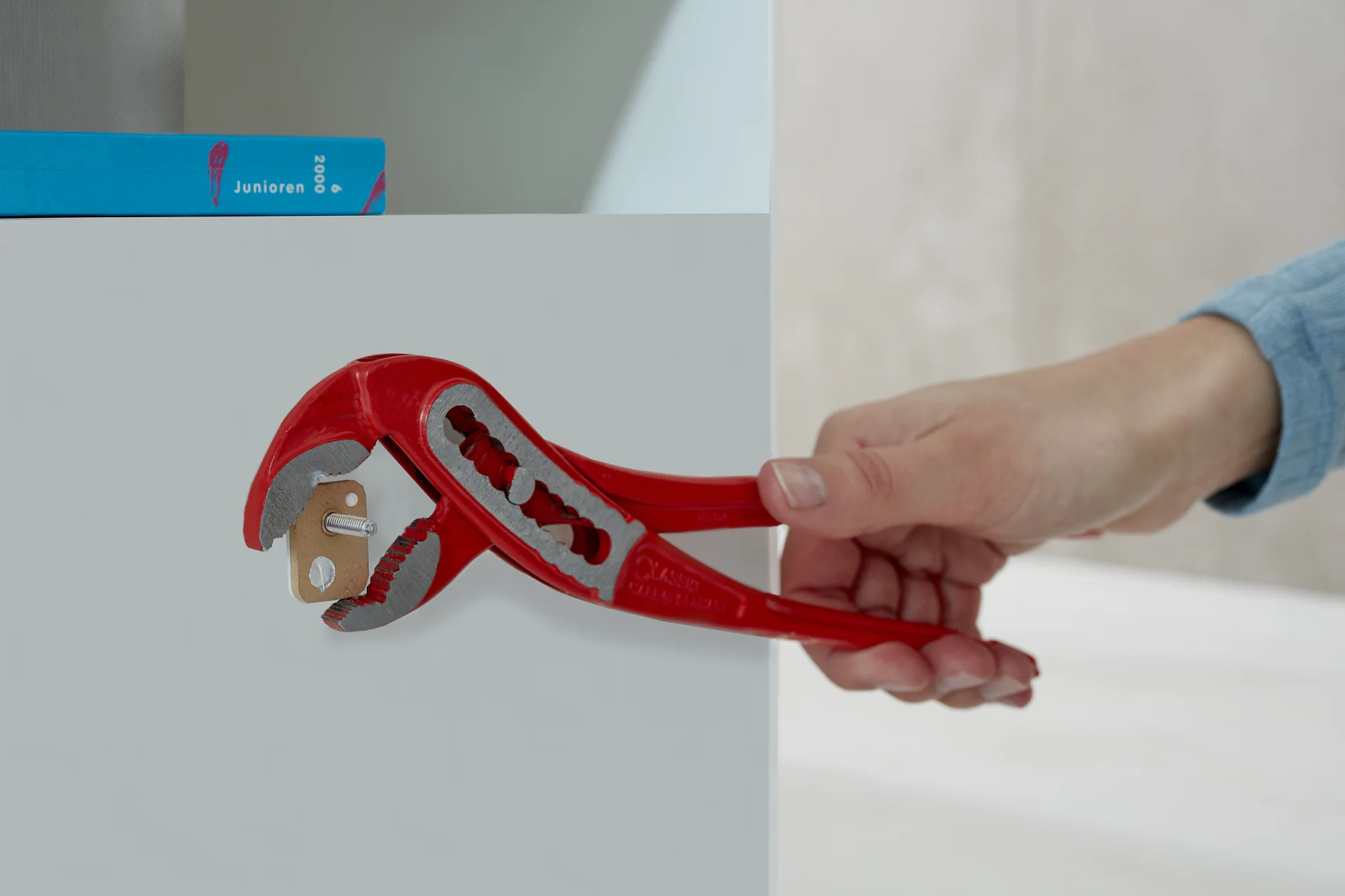 A hand holds a red adjustable wrench gripping a key, which is inserted into a lock on a gray surface with tesa tape. A blue book is partially visible in the background. (This text has been generated by AI)