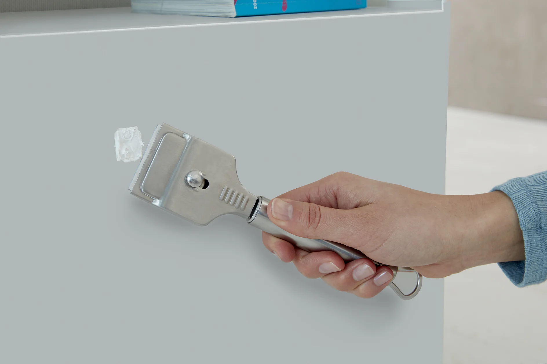 A person uses a metal scraper to remove a white tesa tape adhesive spot from a light gray surface. The person is holding the scraper with their right hand. Part of a book is visible on top of the surface in the background. (This text has been generated by AI)