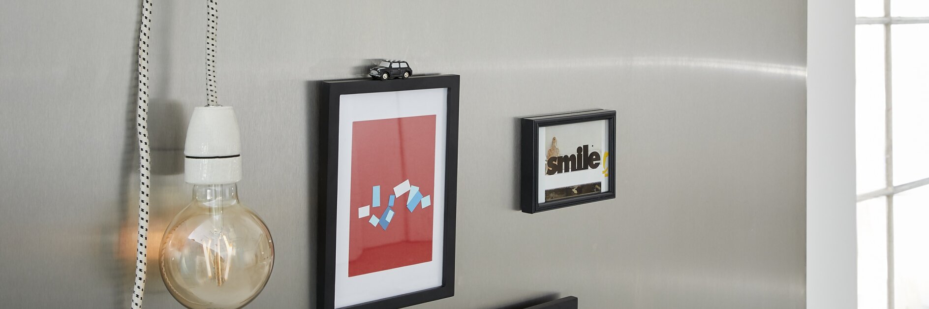A metallic wall features a hanging light bulb on a chain and three framed pictures. The pictures include abstract designs and one with the word smile. In the background, there are books on a shelf near a window, all securely placed using tesa tape. (This text has been generated by AI)