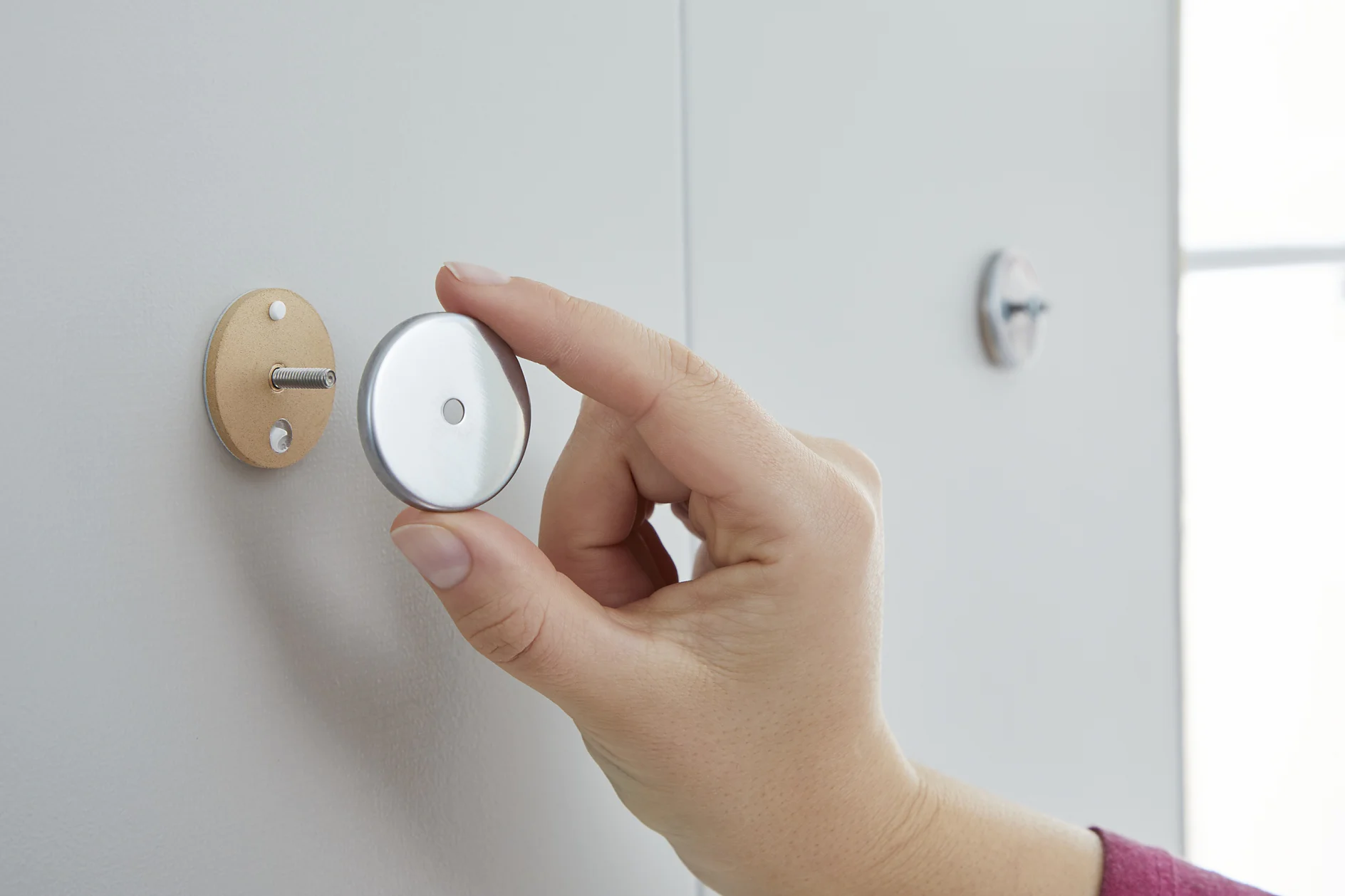A hand is holding a round, metallic wall mount with a screw and a small circular piece. The mount appears to be used for securing objects to a wall using tesa tape. Another similar piece is already attached to the white wall with tesa tape. (This text has been generated by AI)