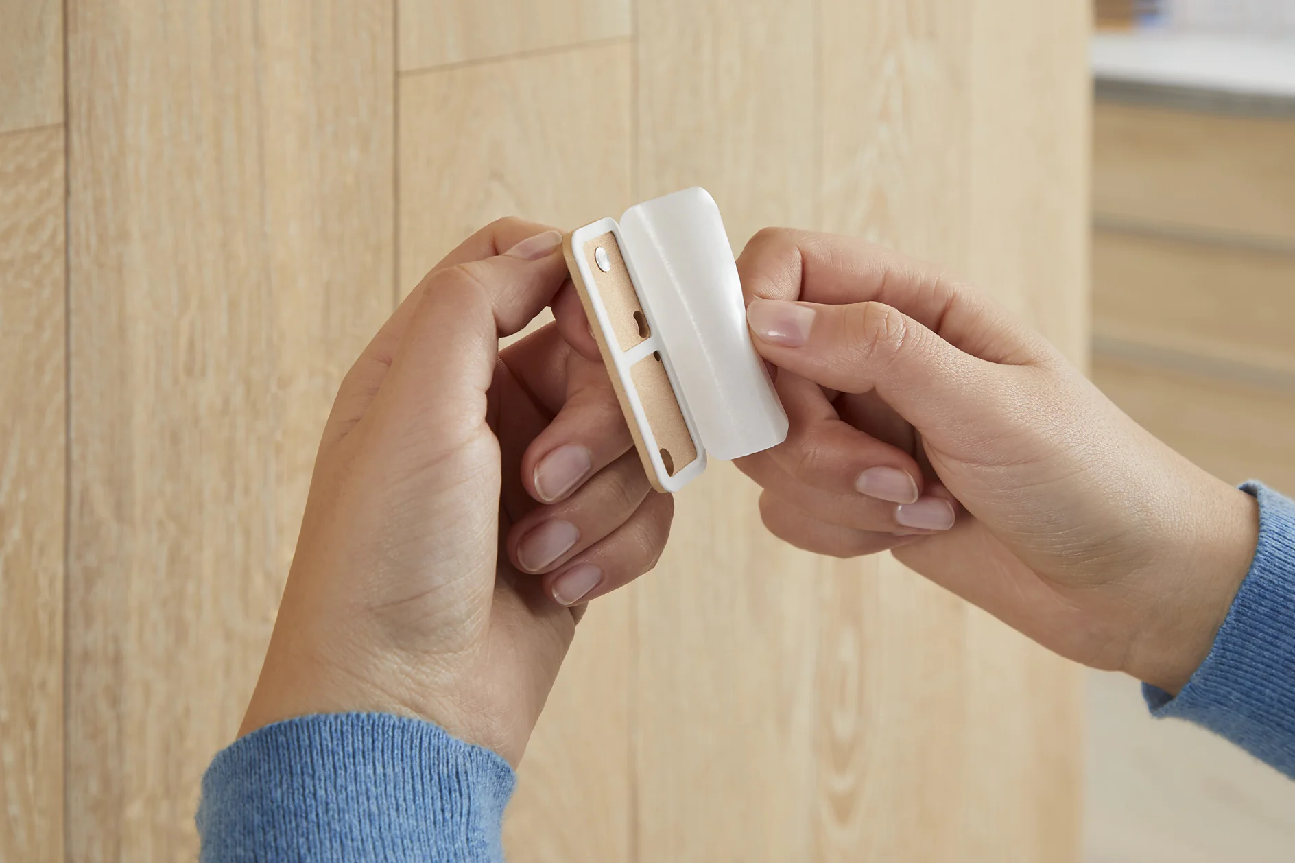 Hands holding a small white plastic device with a wooden backing. The device appears to be a wall-mounted holder or bracket secured with tesa tape. The background is a wooden wall. (This text has been generated by AI)