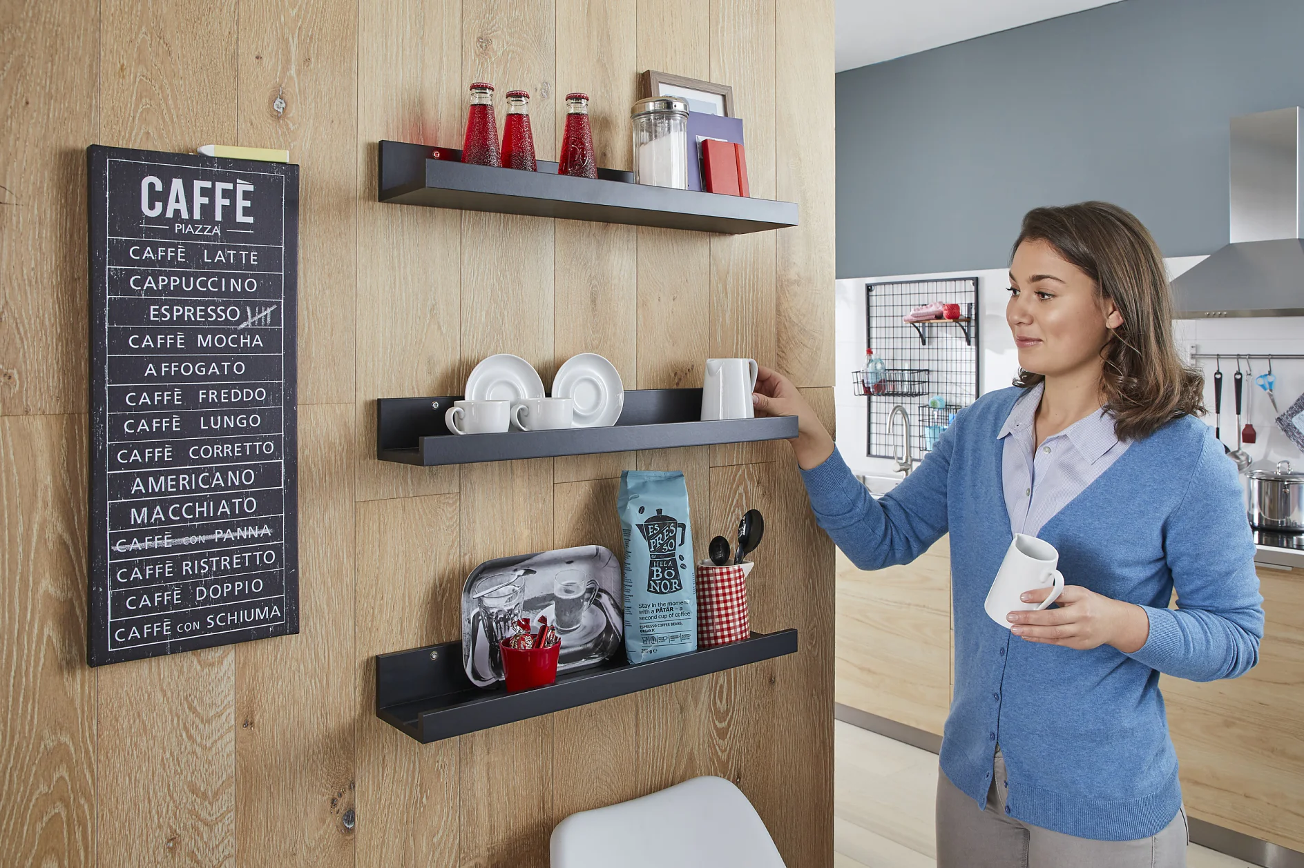 A woman in a blue cardigan places a mug on black wall shelves decorated with coffee items. A menu board displaying coffee types hangs on the wooden wall, secured with tesa tape. The kitchen in the background features modern decor and a red stool. (This text has been generated by AI)