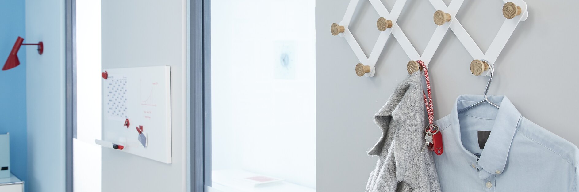 A white accordion coat rack with several pegs is mounted on a light gray wall. A gray coat and a light blue shirt are hanging on it. In the background, there is a tesa tape whiteboard and a red wall hook. (This text has been generated by AI)
