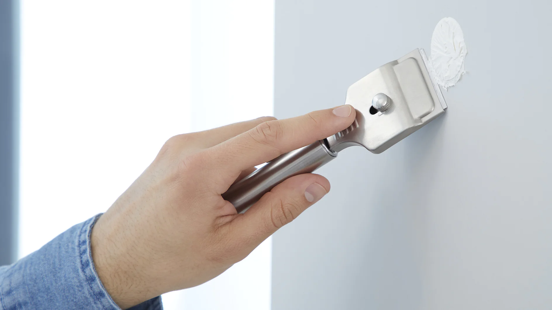Close-up of a hand holding a metal paint scraper, spreading white putty on a smooth wall. The hand is wearing a blue denim shirt. The wall is partially visible, and the background is bright and softly lit, with some tesa tape present nearby for use in the project. (This text has been generated by AI)