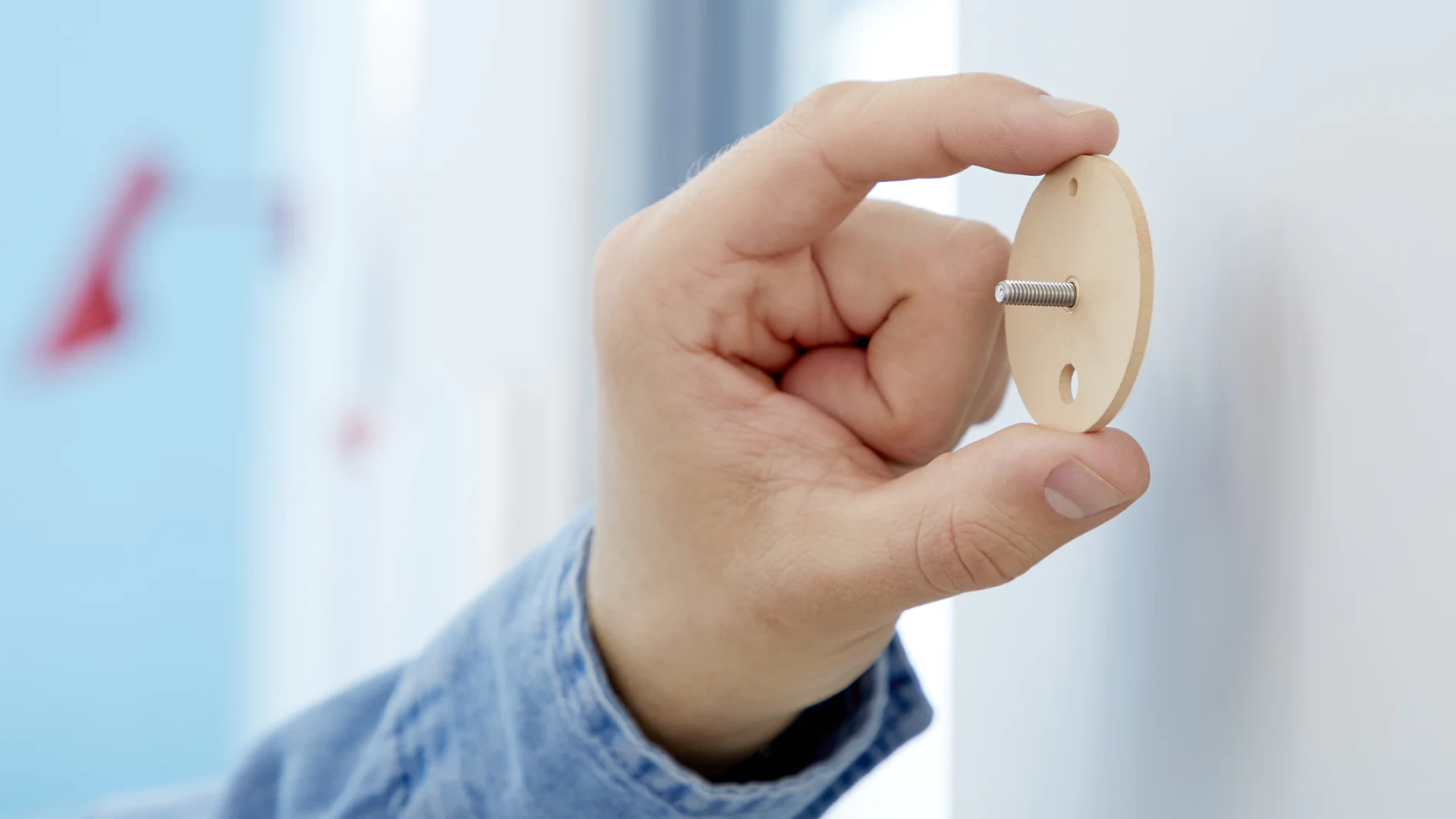 A hand is holding a round beige object featuring a short metallic rod protruding from its center. The background is slightly blurred, showcasing a hint of red on the left within a blue and white color scheme. (This text has been generated by AI)