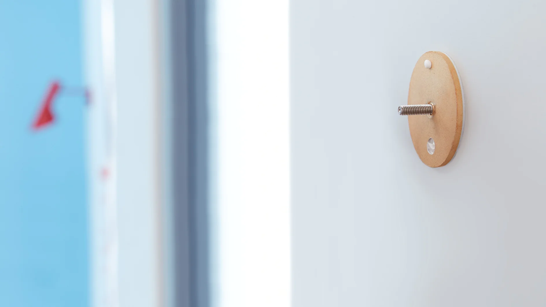 Close-up of a light-colored wooden base with a metal screw protruding from it, attached to a white surface using tesa tape. The background shows a blurred view of a blue wall and a red object. (This text has been generated by AI)
