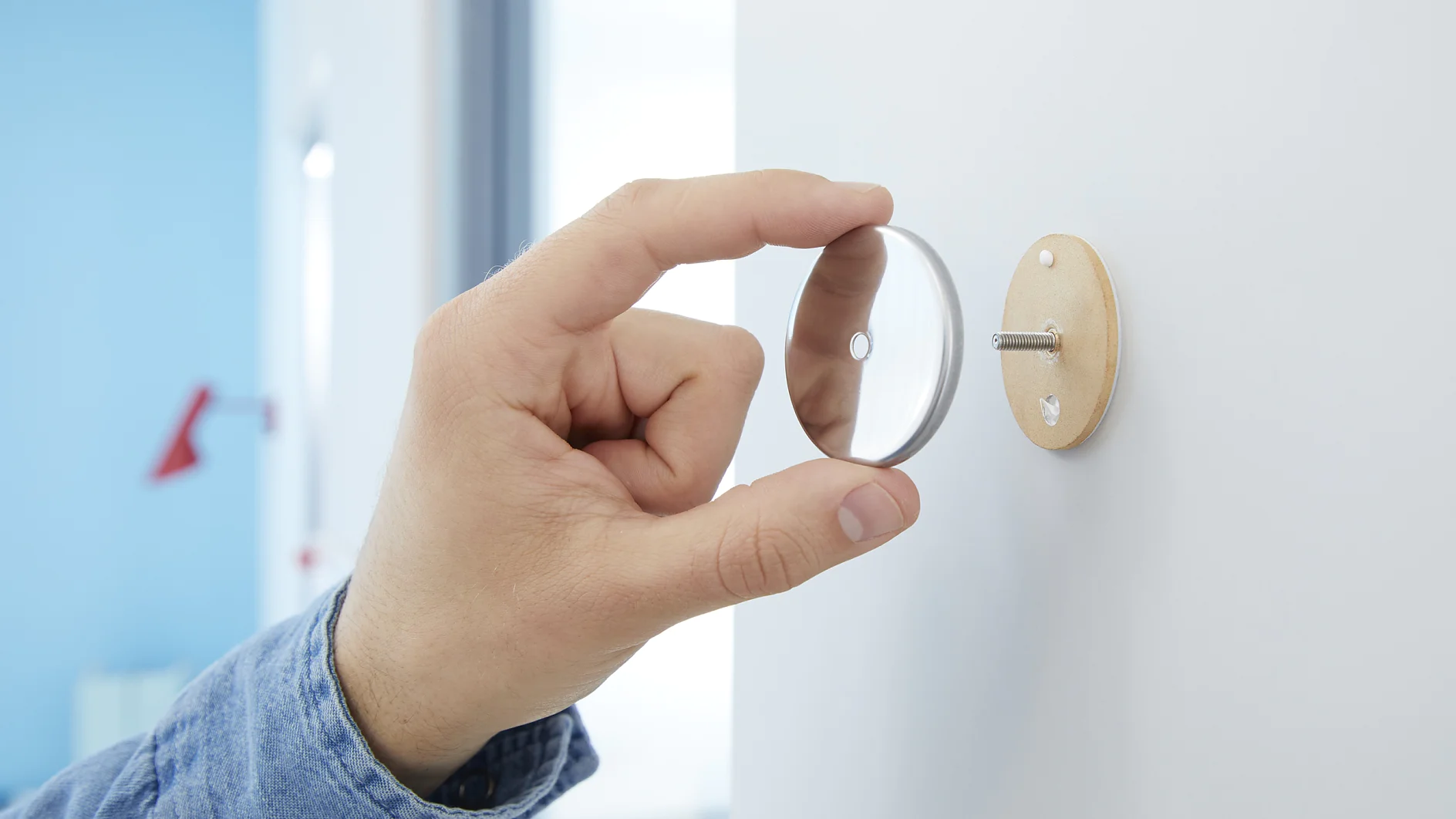 A person is attaching a metal disc to a wall-mounted base. The hand is holding the disc near the base, which is secured with tesa tape and a screw. The background is out of focus, displaying a room with light blue and white walls. (This text has been generated by AI)