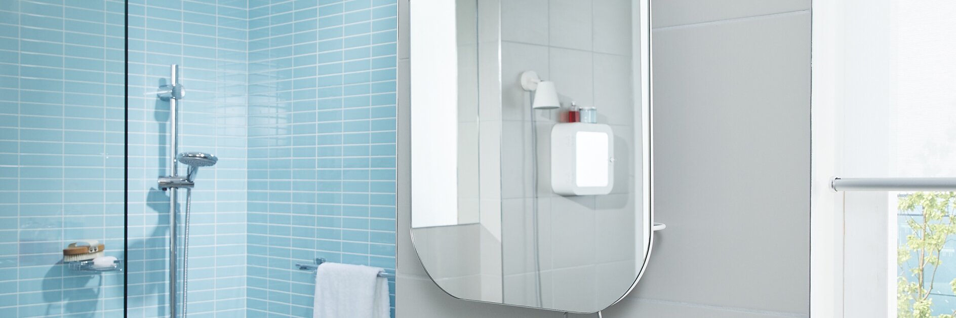 A modern bathroom features light blue tiled walls and a glass shower enclosure. A rounded rectangular mirror is mounted on a gray wall, secured in place with tesa tape. A towel hangs below the mirror. Sunlight filters in through a window on the right. (This text has been generated by AI)