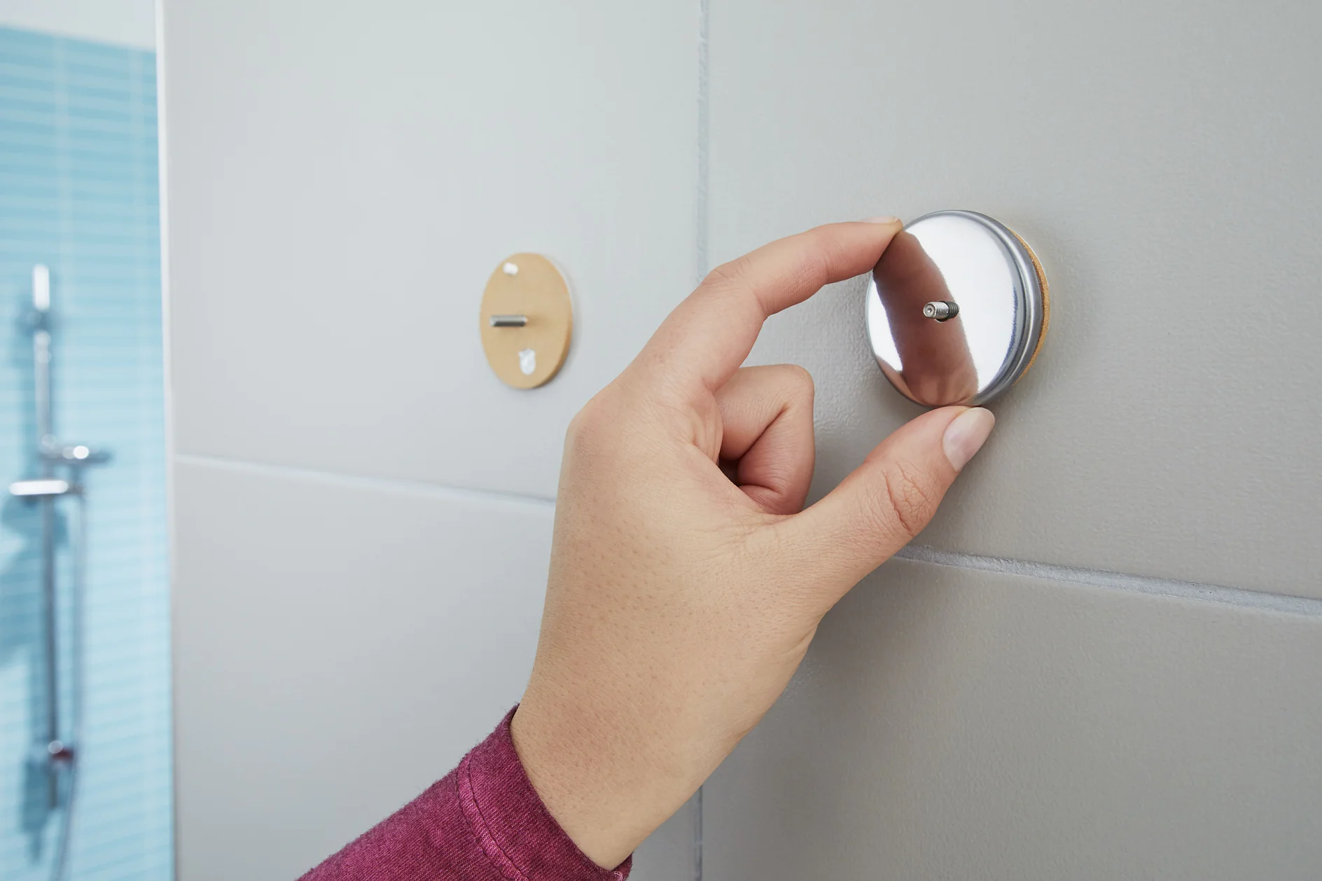 A person is installing a shiny round metal hook onto a gray tiled wall using a screw. Another hook without the metal part is visible in the background. The persons hand shows a sleeve of a mauve shirt. Blue tiles are seen on the left. (This text has been generated by AI)