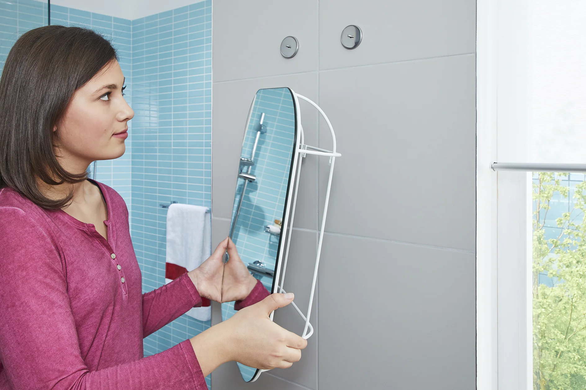 A woman in a pink top is positioning a round mirror on a bathroom wall with blue tiles using tesa tape. The mirror has a white frame. A towel is hanging on a rail in the background, and sunlight streams through a window. (This text has been generated by AI)