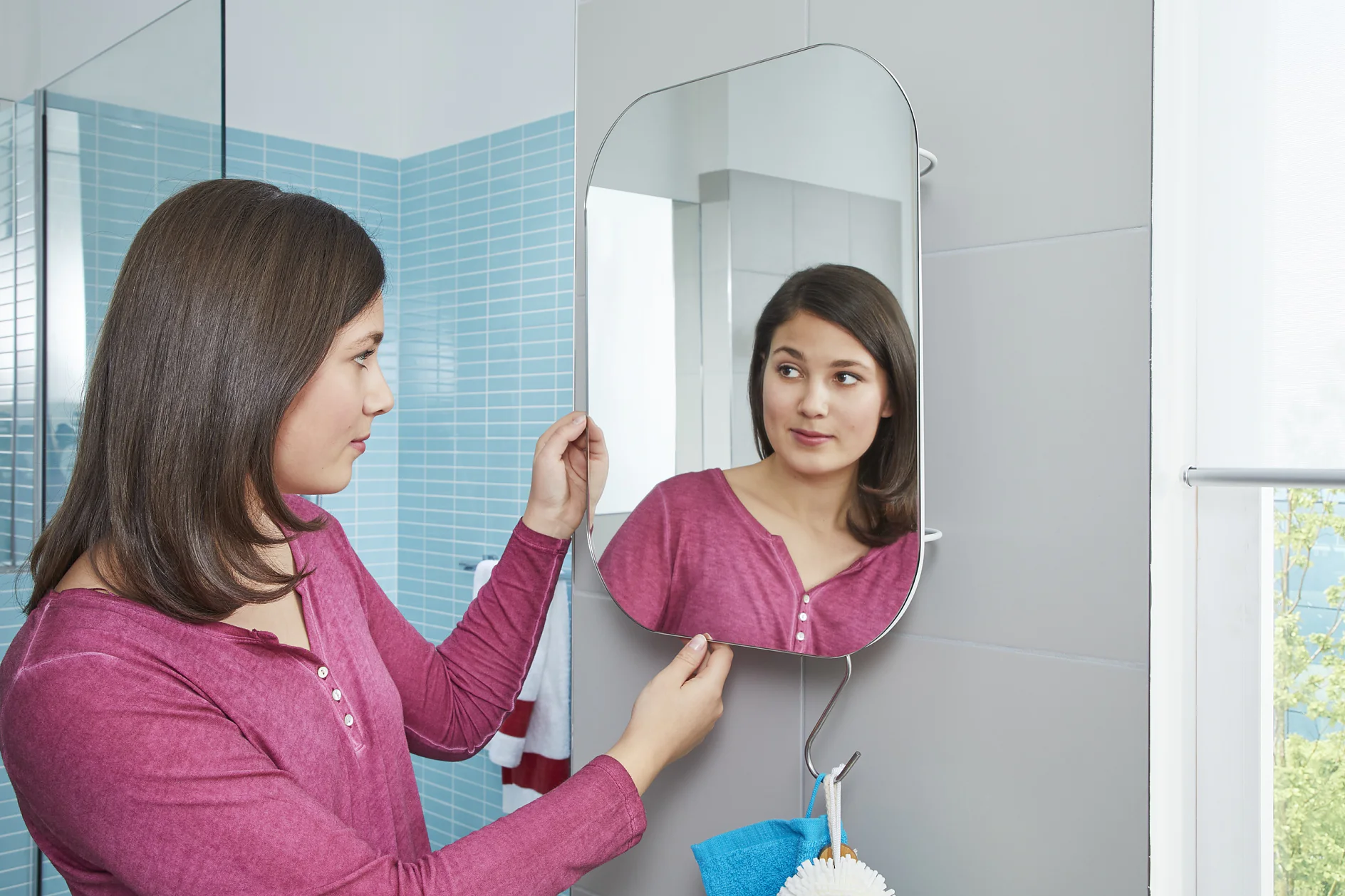 A woman wearing a pink long-sleeve shirt looks at her reflection in a bathroom mirror. The walls are tiled in blue, and a small towel hangs below the mirror secured with tesa tape. Natural light enters through a window on the right. (This text has been generated by AI)