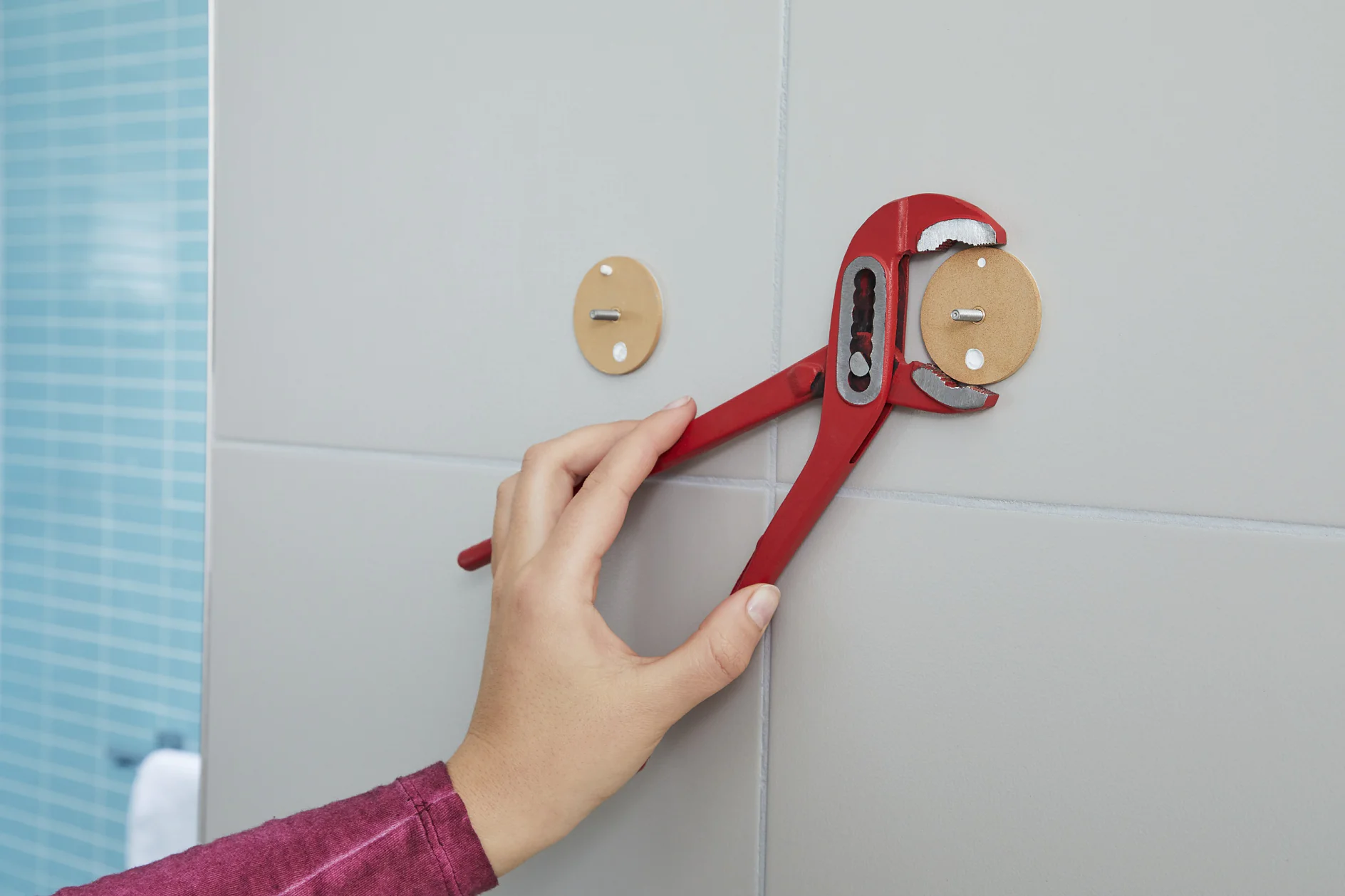 A person wearing a maroon sleeve uses a red pipe wrench to install or adjust a circular fitting on a tiled wall. The wall has a beige fitting with two visible screws. Blue tiles are partially visible in the background, with some sections secured using tesa tape. (This text has been generated by AI)