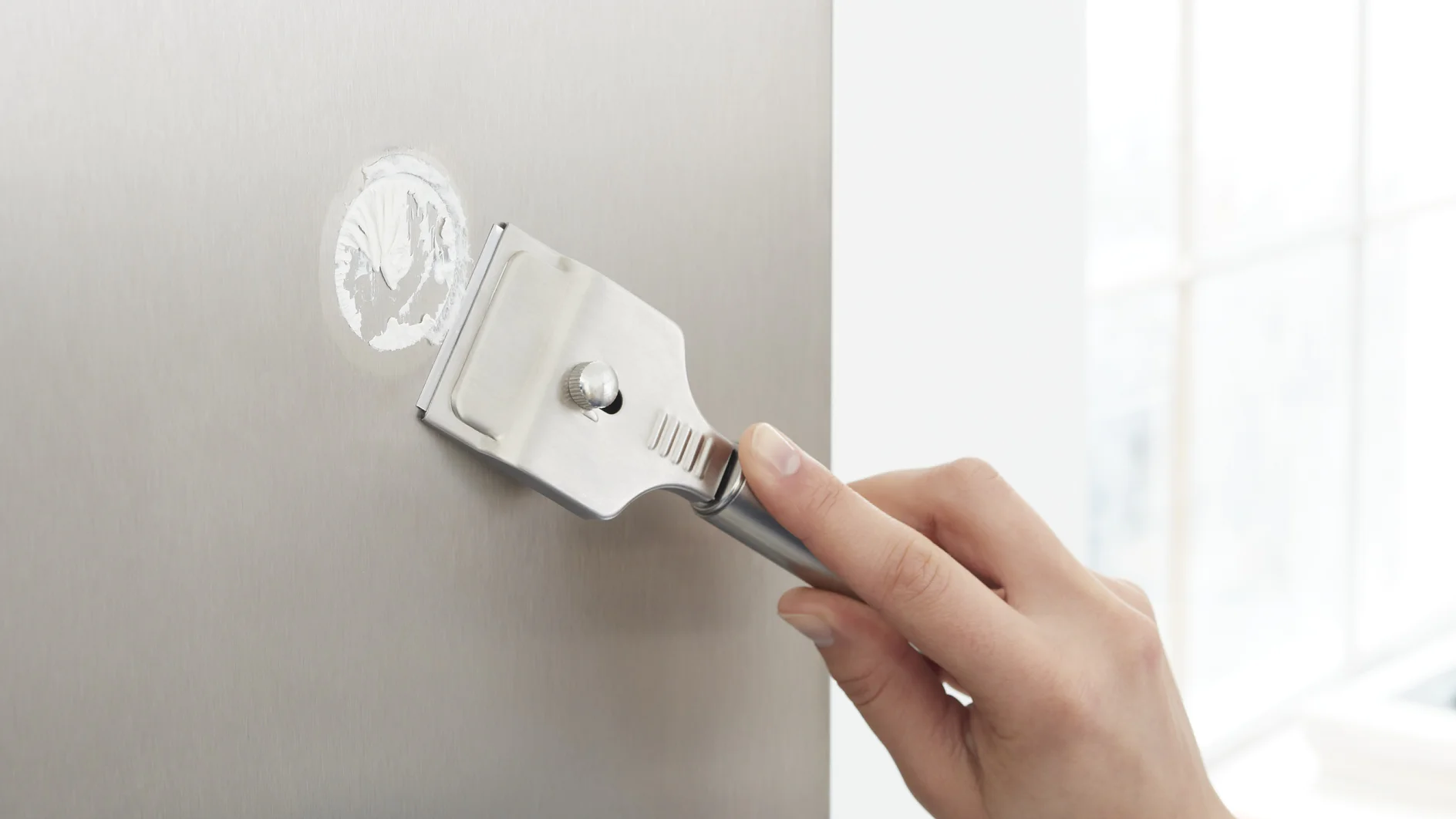 A person is using a metal scraper to remove a circular tesa tape residue from a flat, shiny surface. The background is blurred and bright, suggesting natural light from a nearby window. (This text has been generated by AI)