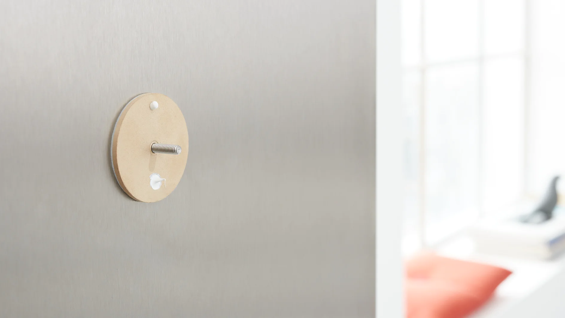 A metallic wall with a beige, circular piece attached, featuring a central rod. A small amount of white tesa tape is visible around the piece. The background shows a softly focused window with light coming in, revealing a blurred red cushion. (This text has been generated by AI)